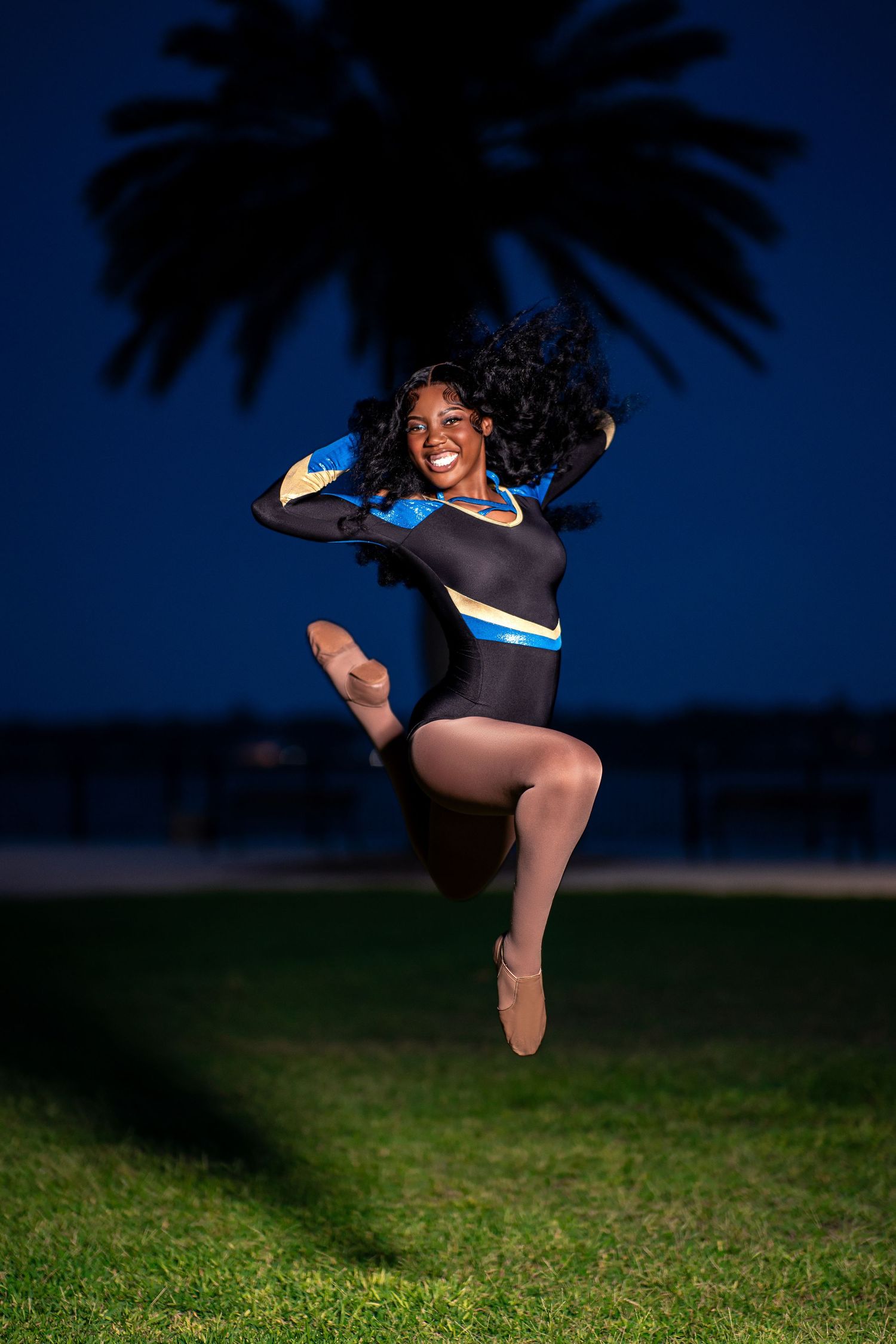 Cheerleader in blue and black uniform jumping joyfully near palm tree silhouette at night.