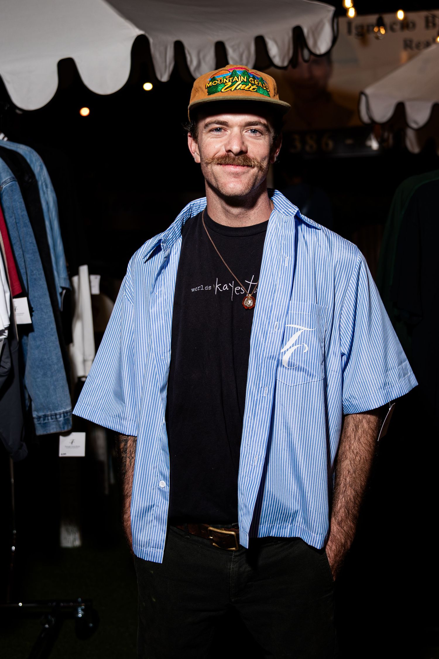 Smiling man in blue striped shirt and orange cap posing at outdoor Christian apparel event.