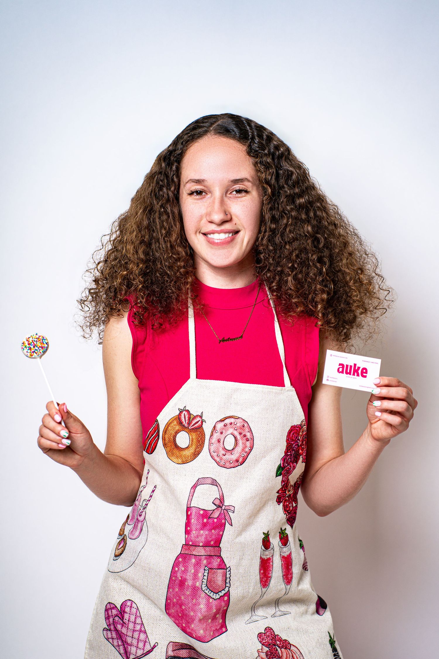 Smiling girl in baking apron holding a sprinkle lollipop and Auke business card against white background.