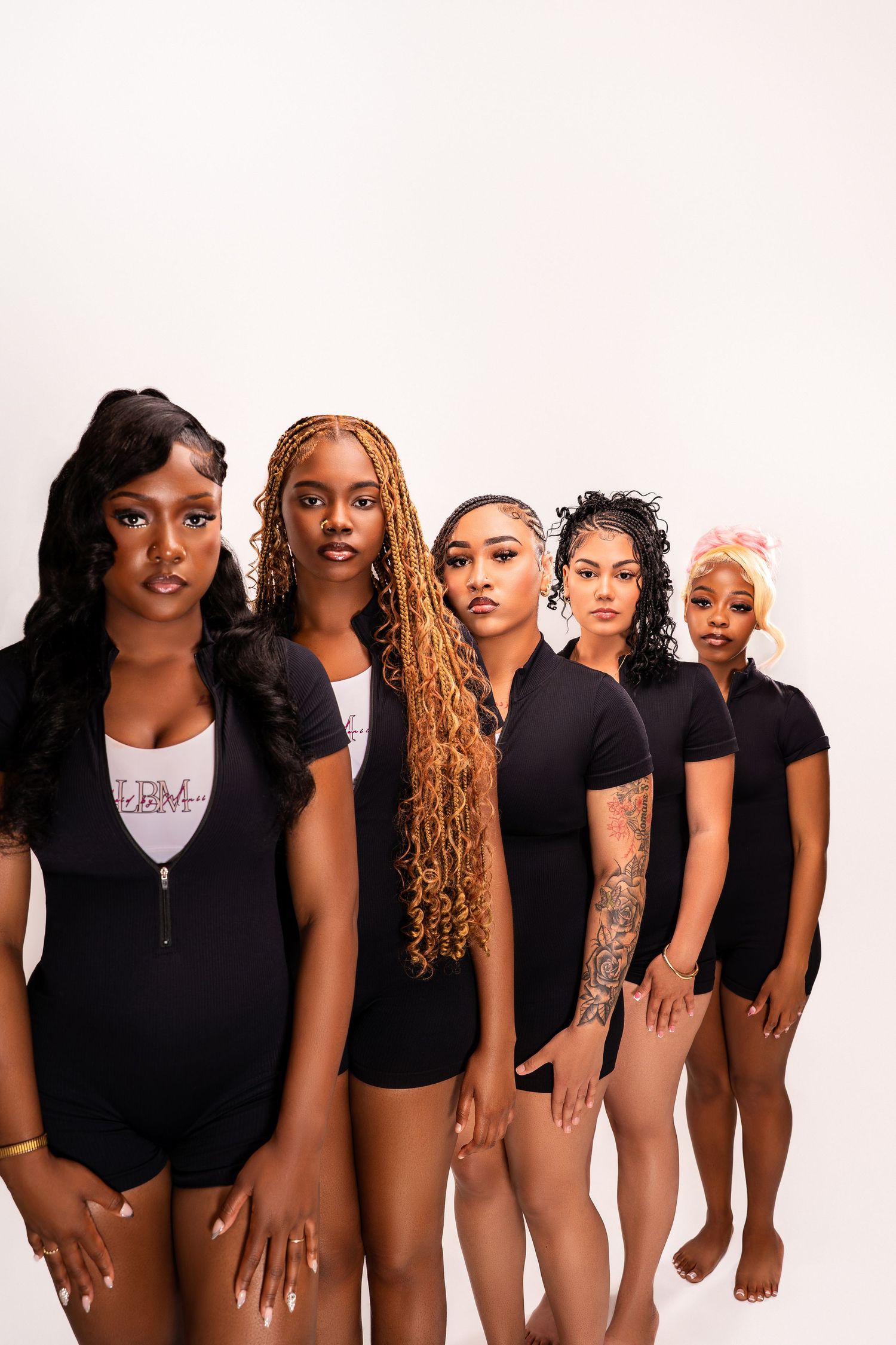 Five young Black women in matching black bodysuits pose confidently in a studio against a white background.