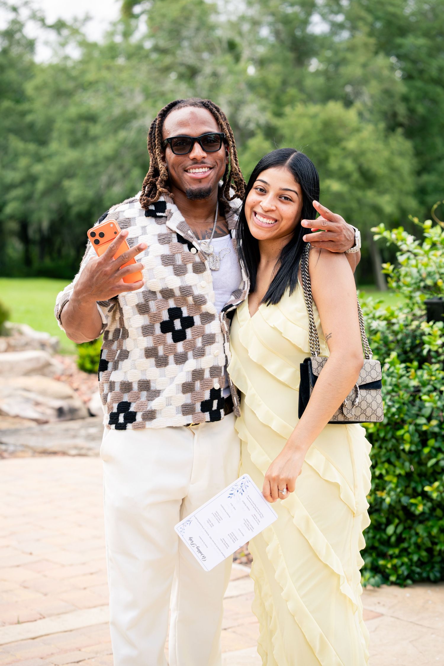 Couple posing outdoors at a wedding, man in patterned shirt holding phone, woman in yellow ruffled dress.