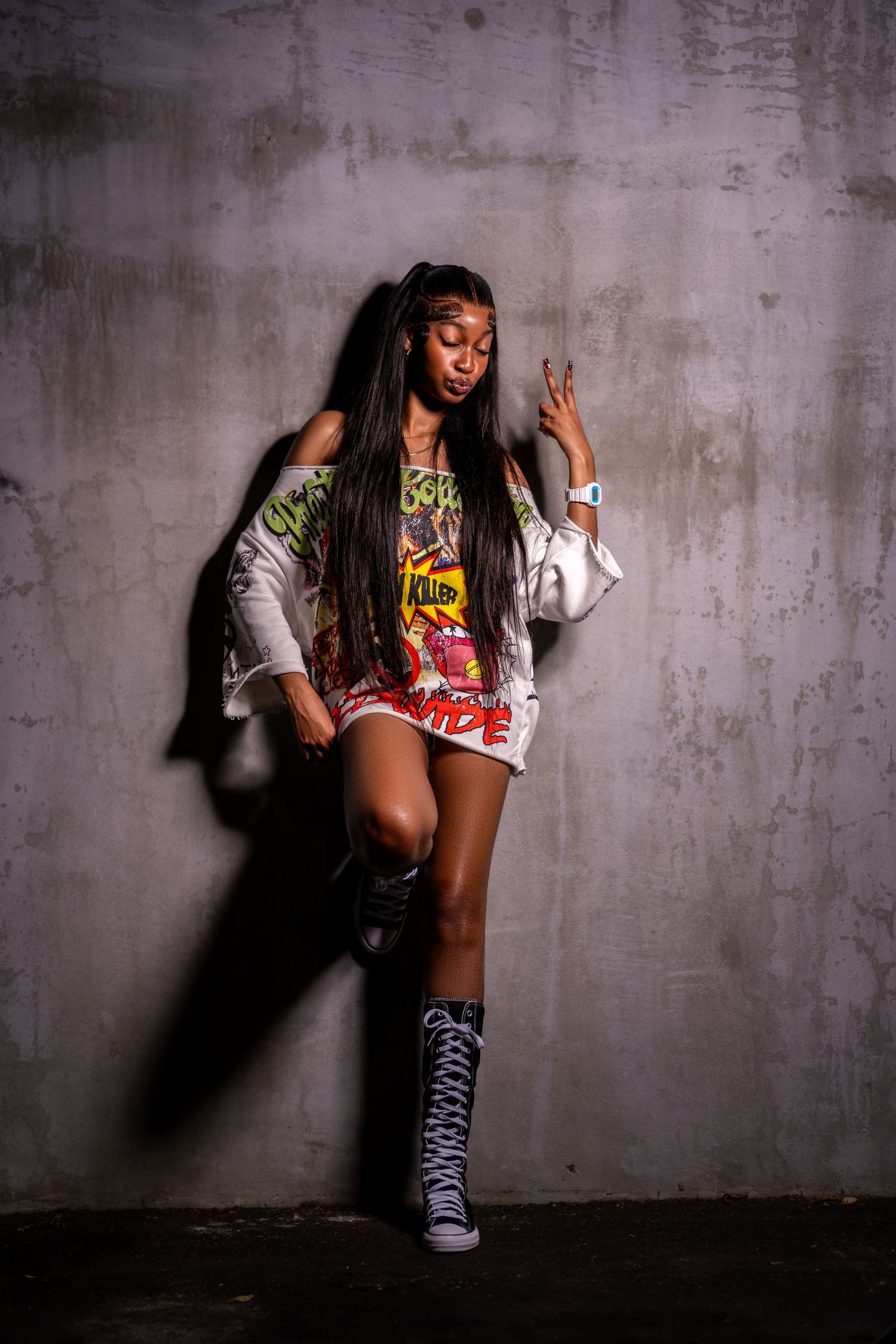 Young Black woman in colorful graphic tee, shorts, and tall boots poses confidently against a concrete wall.