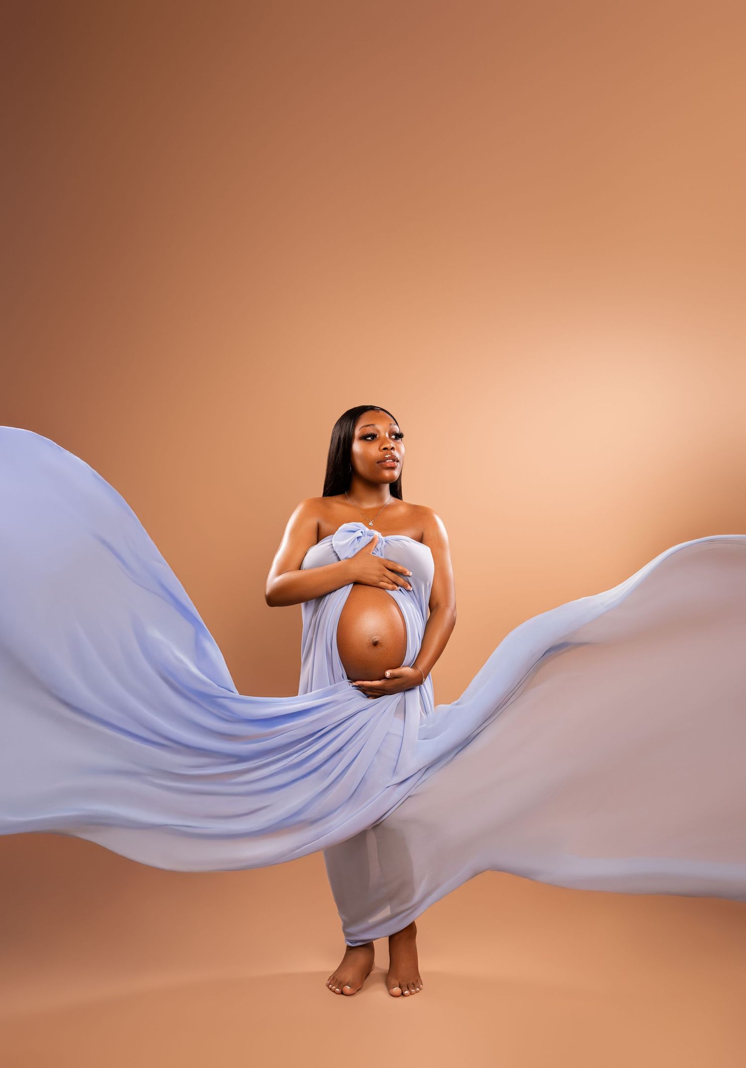 Pregnant woman draped in flowing blue fabric poses gracefully against warm tan background in elegant maternity studio portrait.