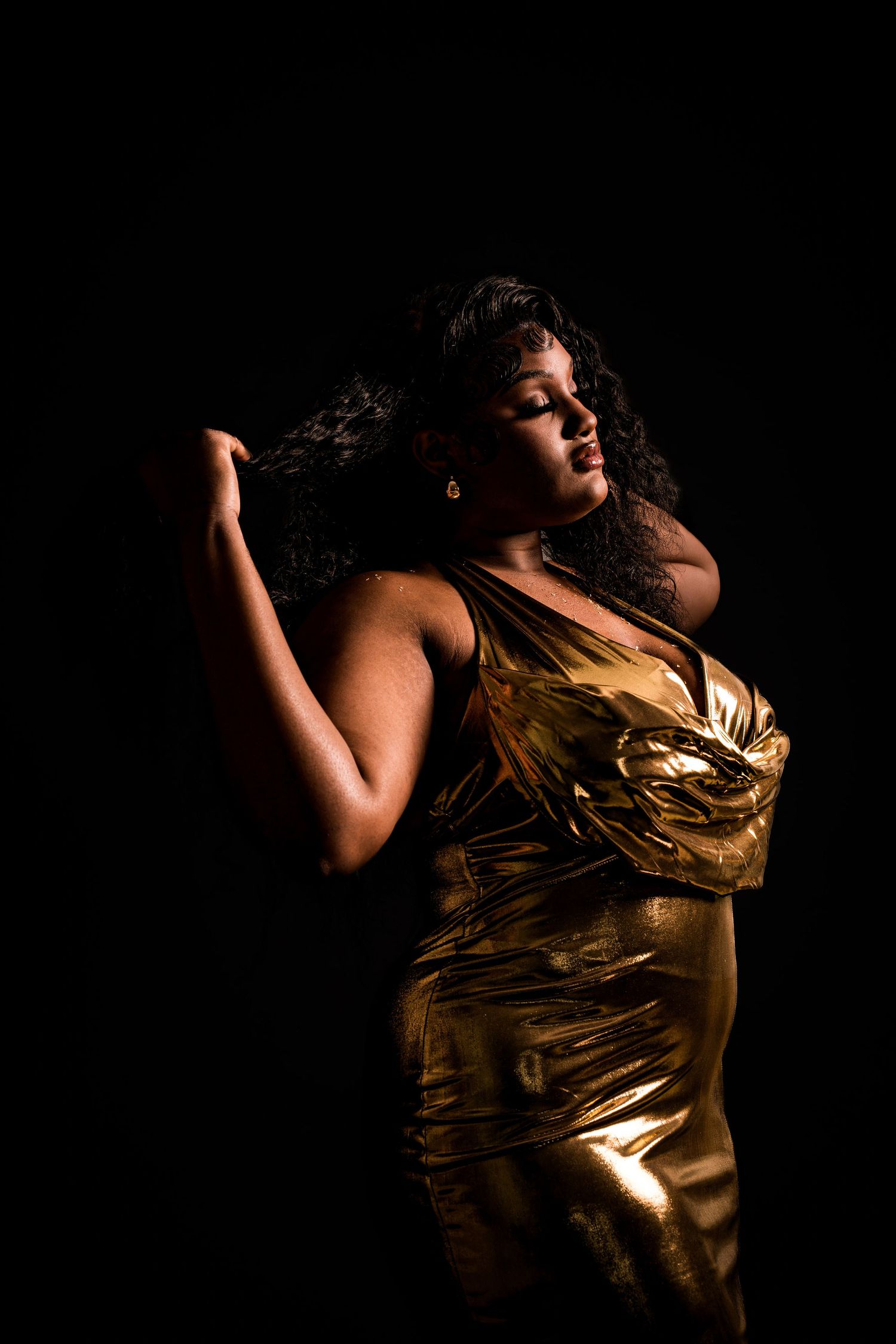 Woman in gold metallic dress posing dramatically against dark black background.