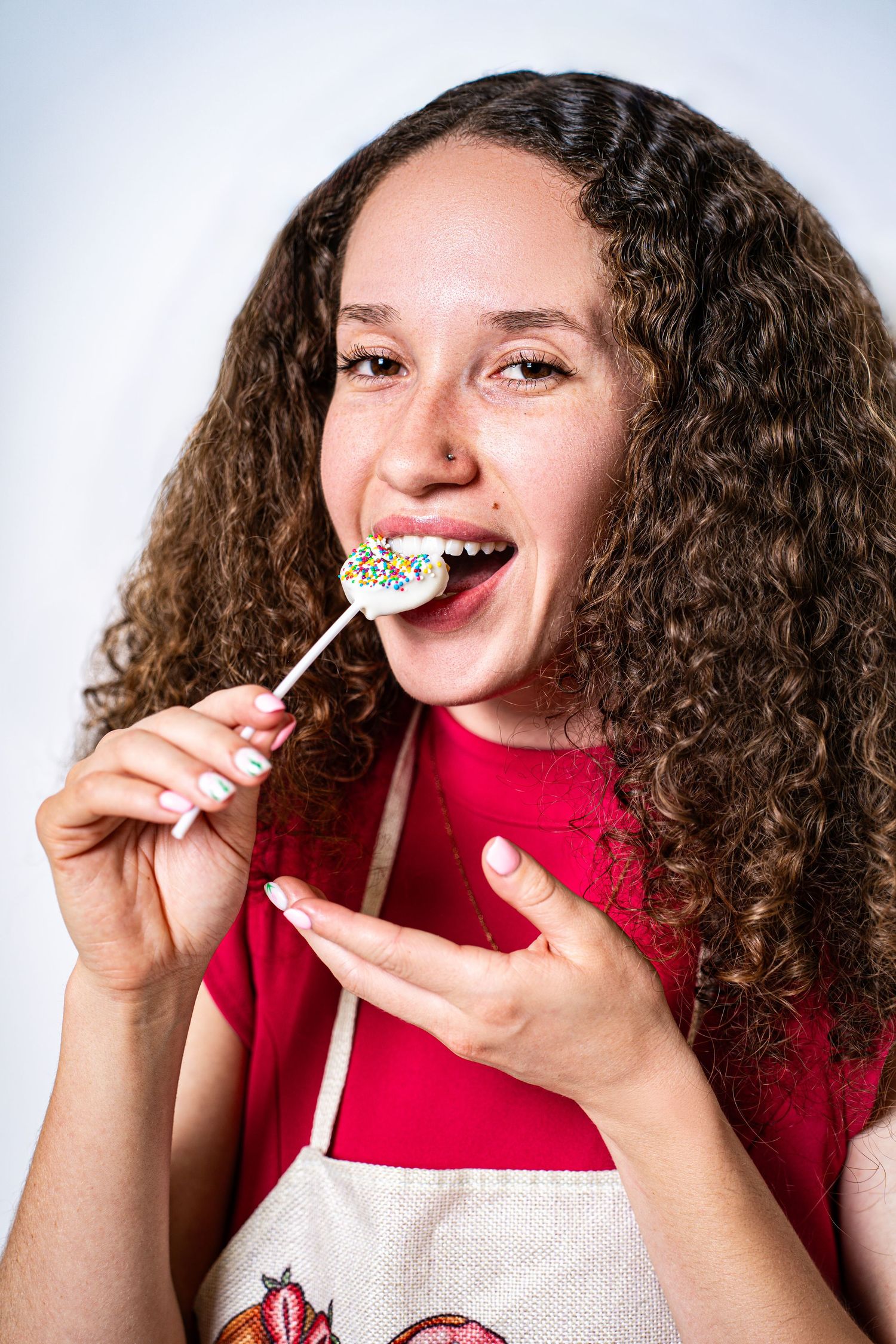 Young woman with curly hair licking a colorful sprinkle lollipop while wearing a red top and baking apron.