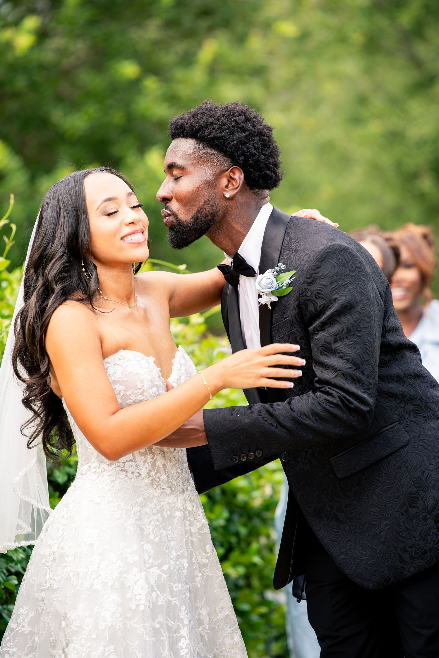 Bride and groom share a tender moment during outdoor wedding ceremony, groom kissing bride's cheek as she smiles.