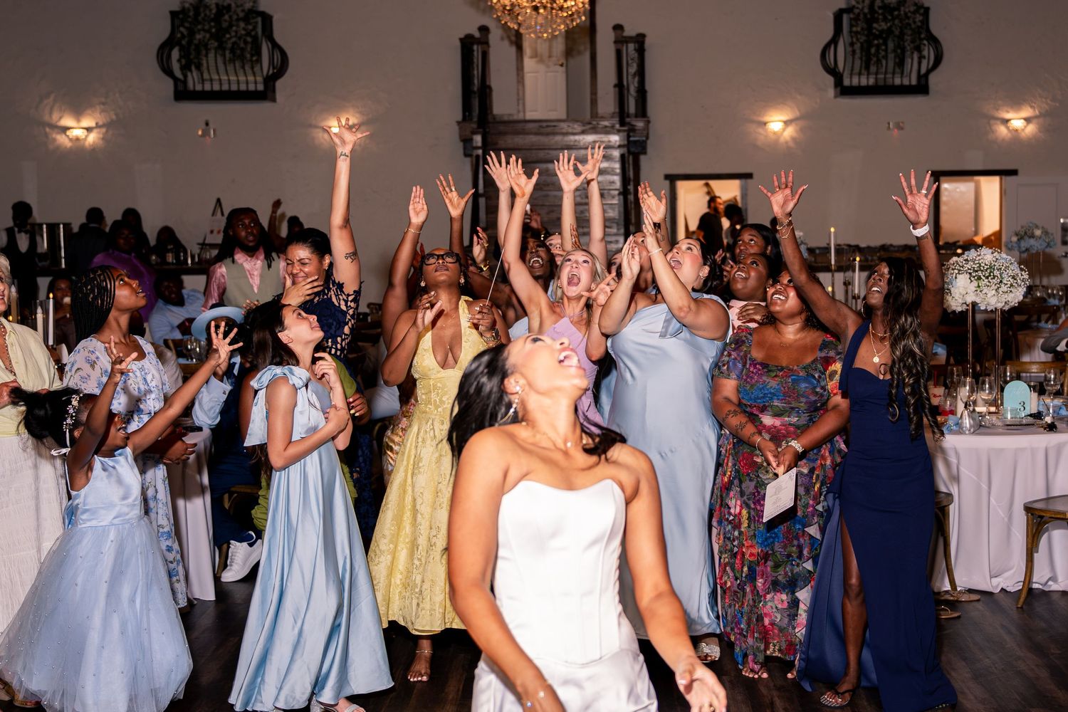 Excited wedding guests reach up to catch bouquet toss during lively indoor wedding reception celebration.
