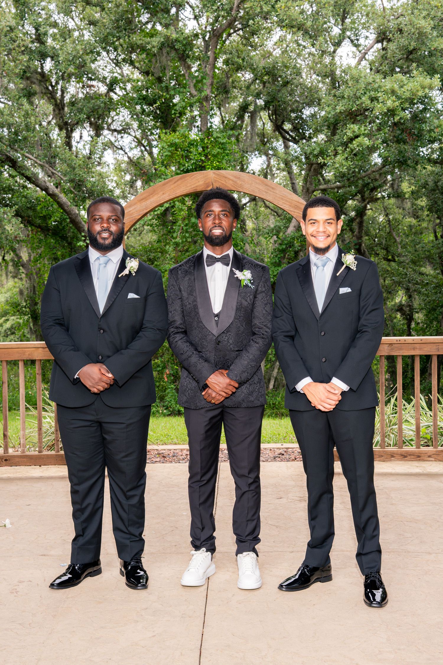 Three groomsmen in black suits pose under wooden arch at outdoor wedding venue.