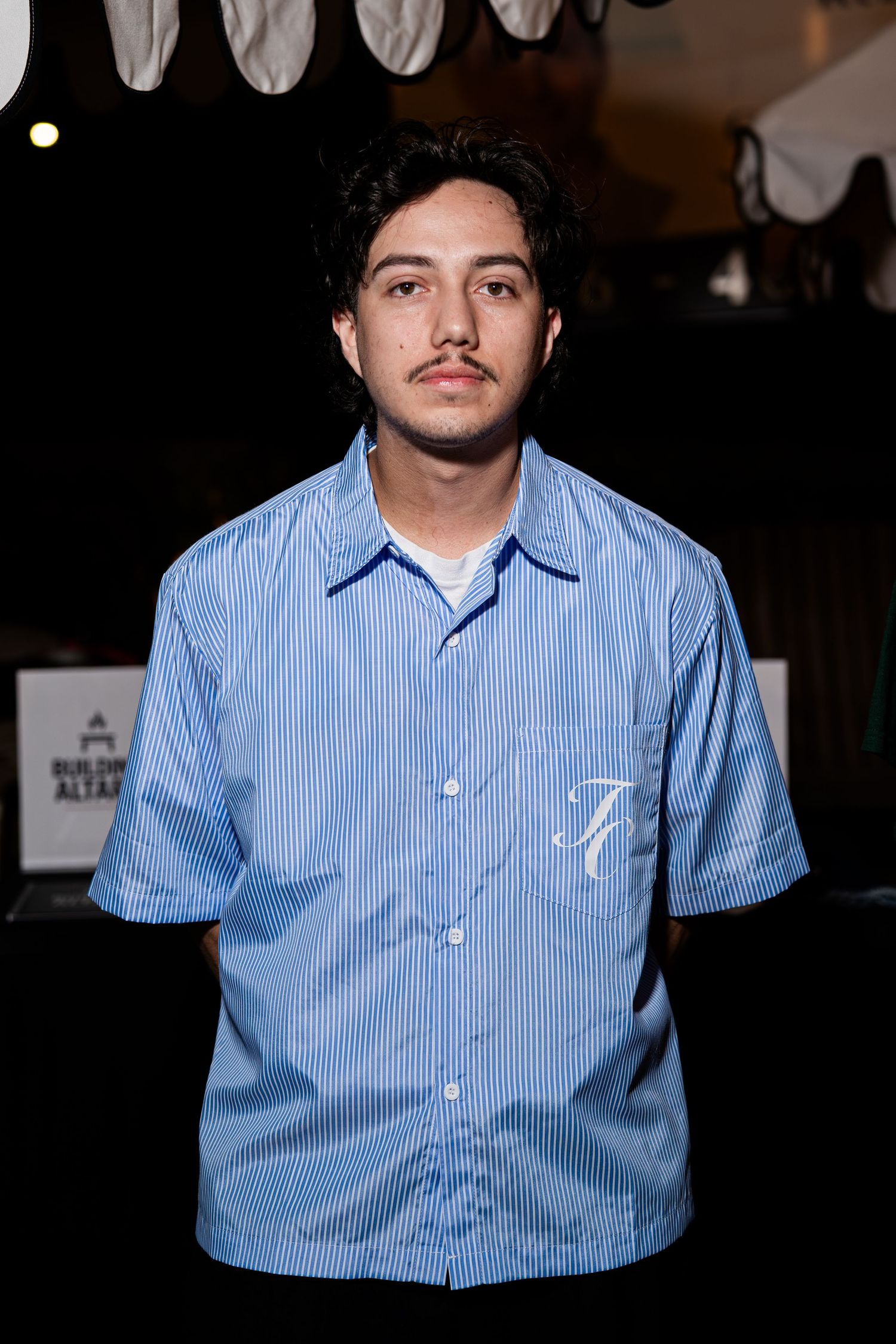 Young man in blue striped short-sleeve button-up shirt with embroidered logo at an indoor event.