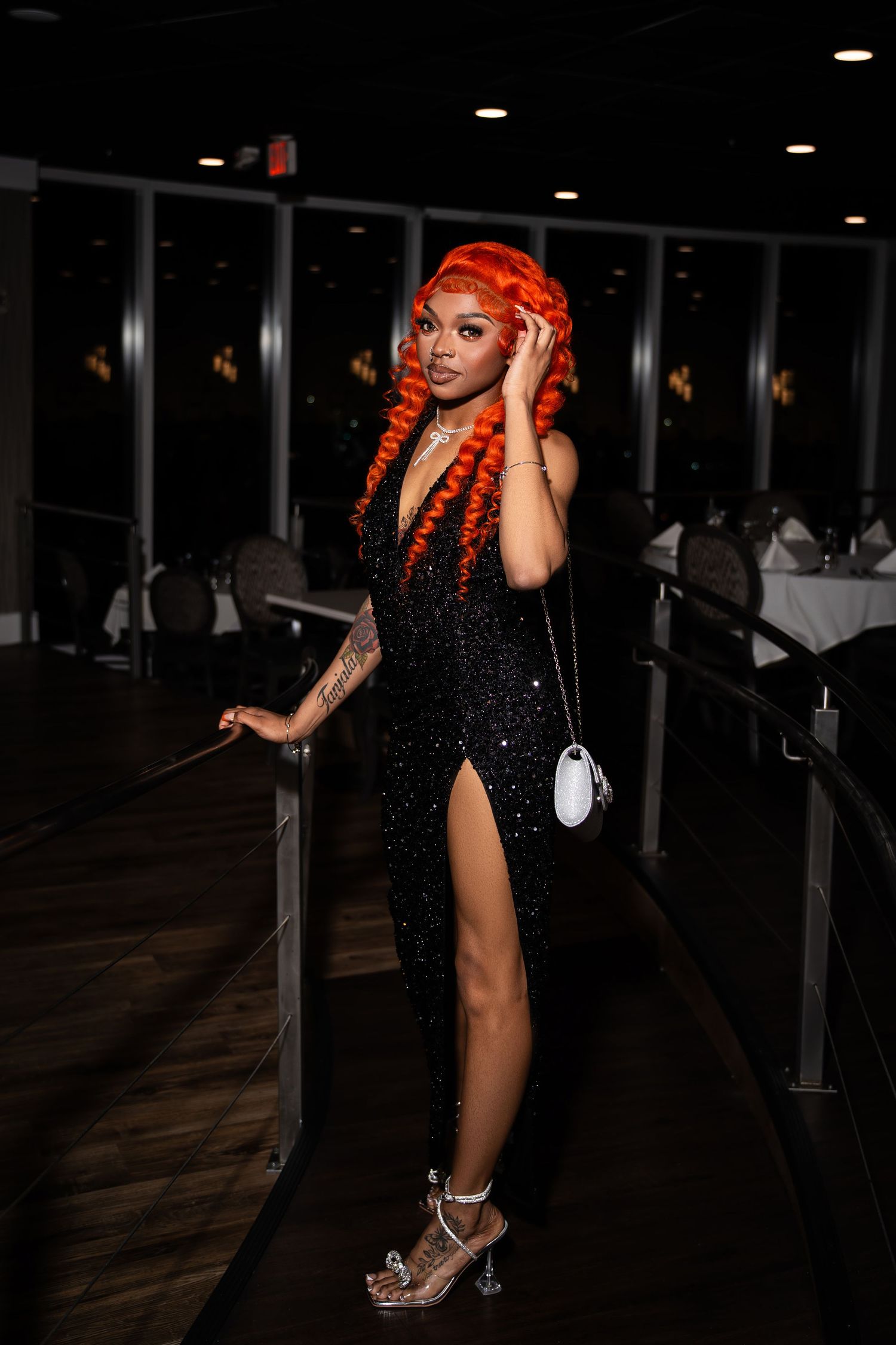 Woman with vibrant orange curly hair wearing black sparkle gown with thigh slit in dark restaurant.