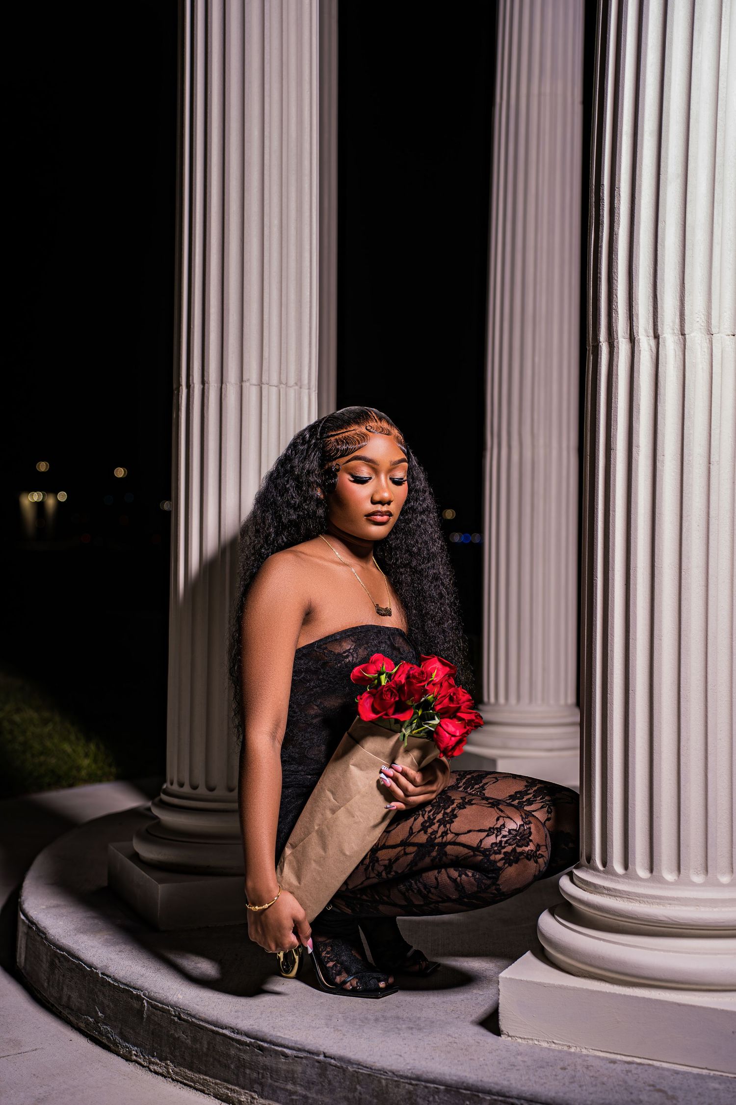 Young Black woman in black lace outfit sits on steps between white columns holding red roses at night.