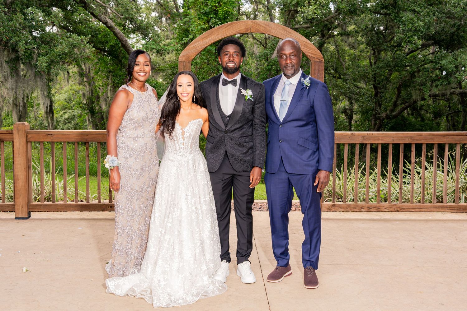 Wedding couple poses with two family members under wooden arch on outdoor venue deck.