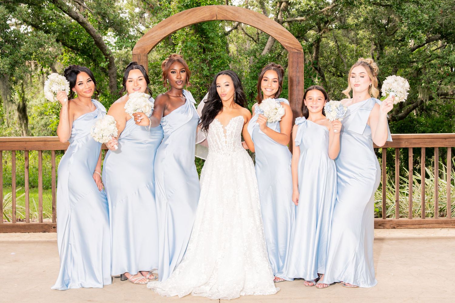 Bride with bridesmaids in light blue dresses holding bouquets under wooden arch outdoors.