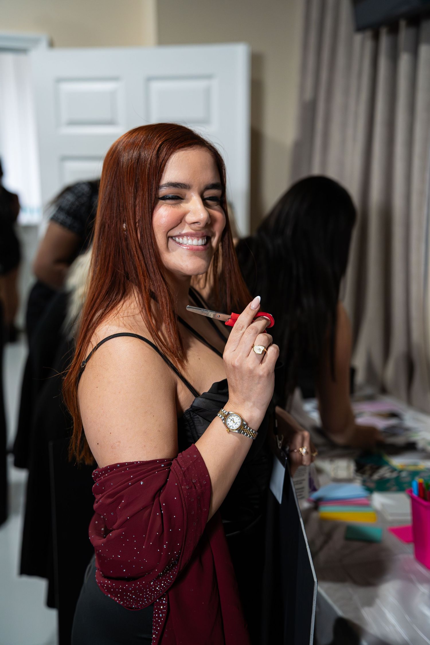 Smiling woman with red hair in burgundy dress holds scissors at event table.