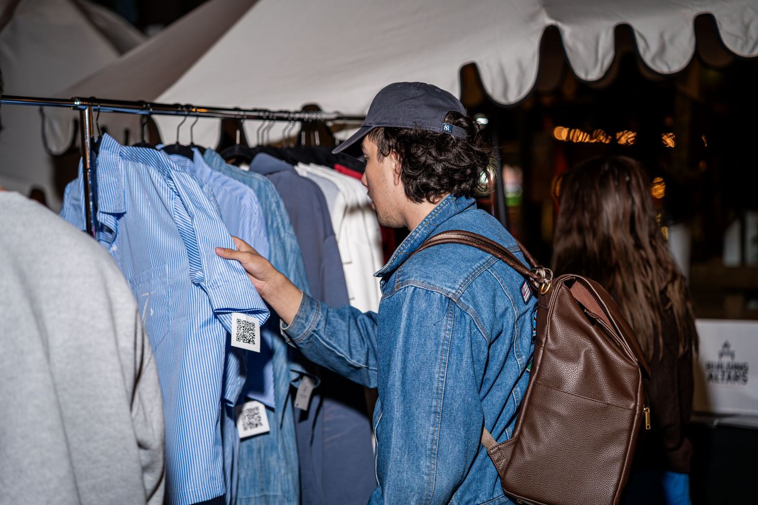 Shoppers browsing blue shirts on a clothing rack at an outdoor retail event at night.