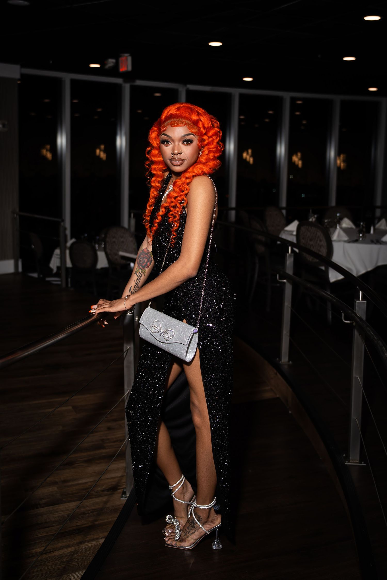 Woman with long orange curly hair in black sequin dress with high slit carries white purse at night.