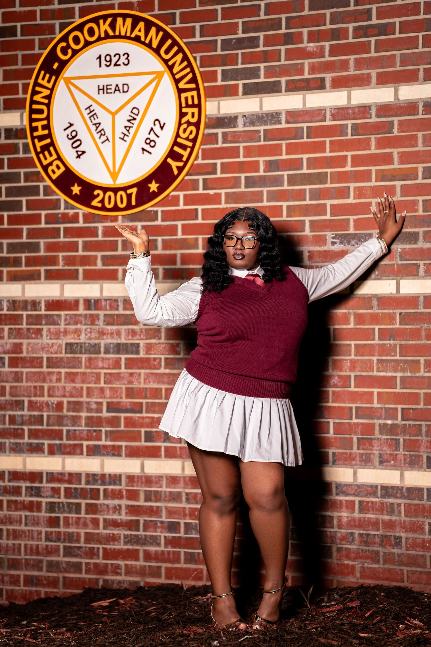 A cheerful Black woman in school-inspired outfit gestures beside Bethune-Cookman University emblem on brick wall.
