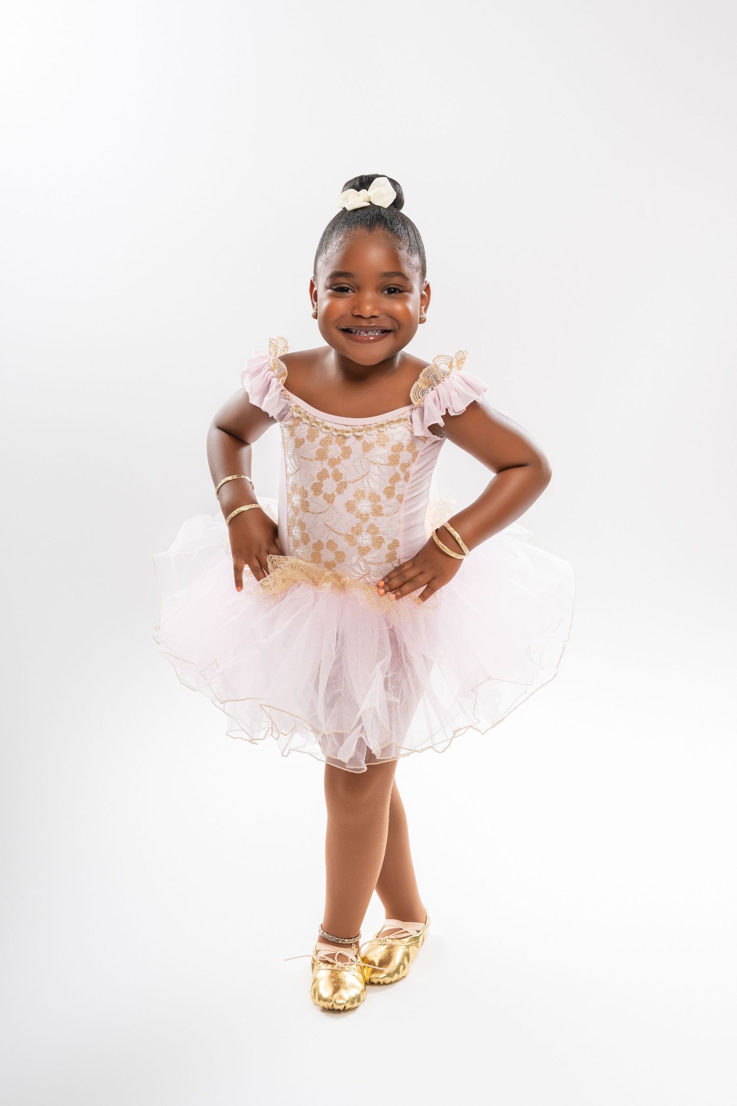 Smiling young Black girl in pink and gold ballet tutu costume with gold shoes posing on white background.