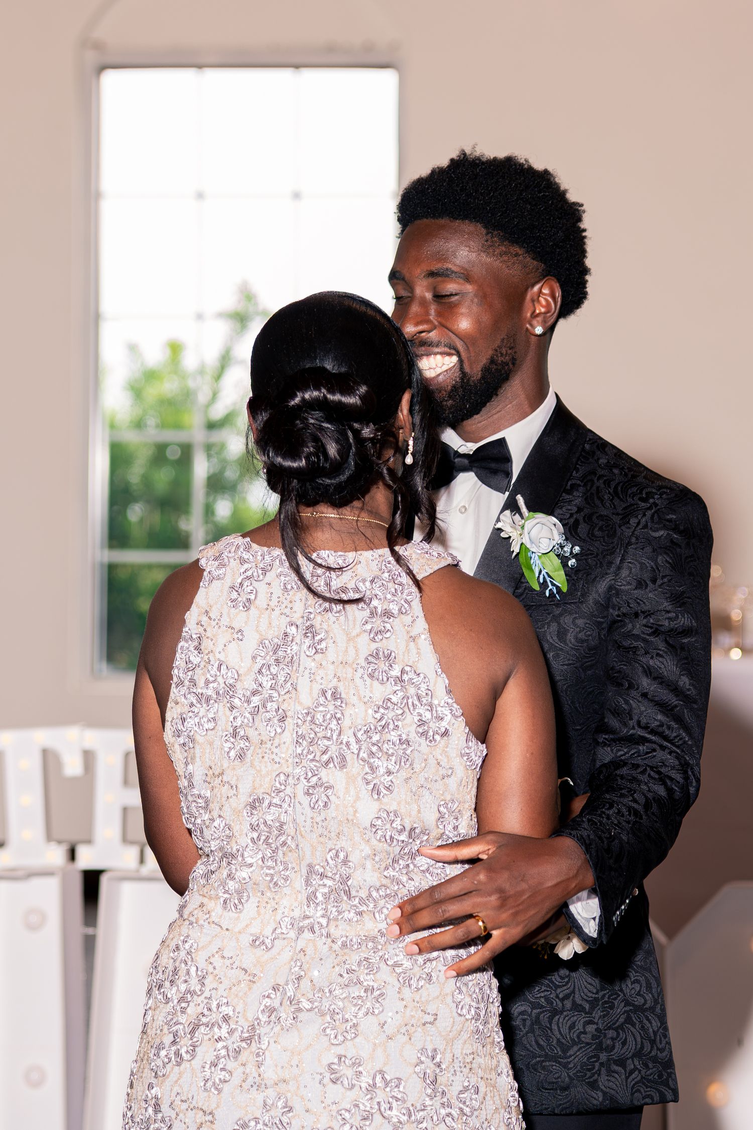 Groom in black tuxedo shares joyful dance with woman in floral dress during wedding reception.