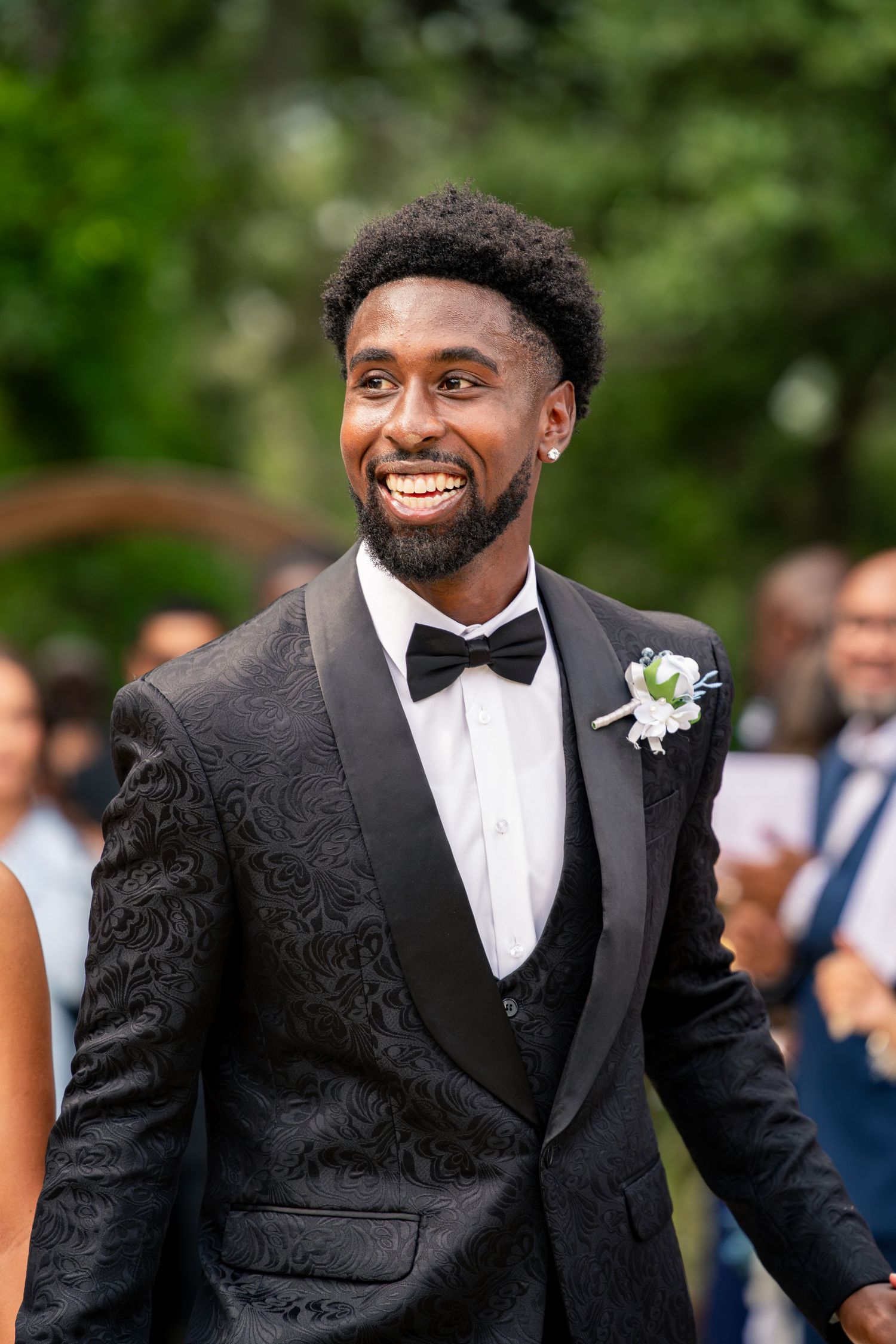 Joyful groom in black floral tuxedo with bow tie and boutonniere smiles broadly at outdoor wedding ceremony.