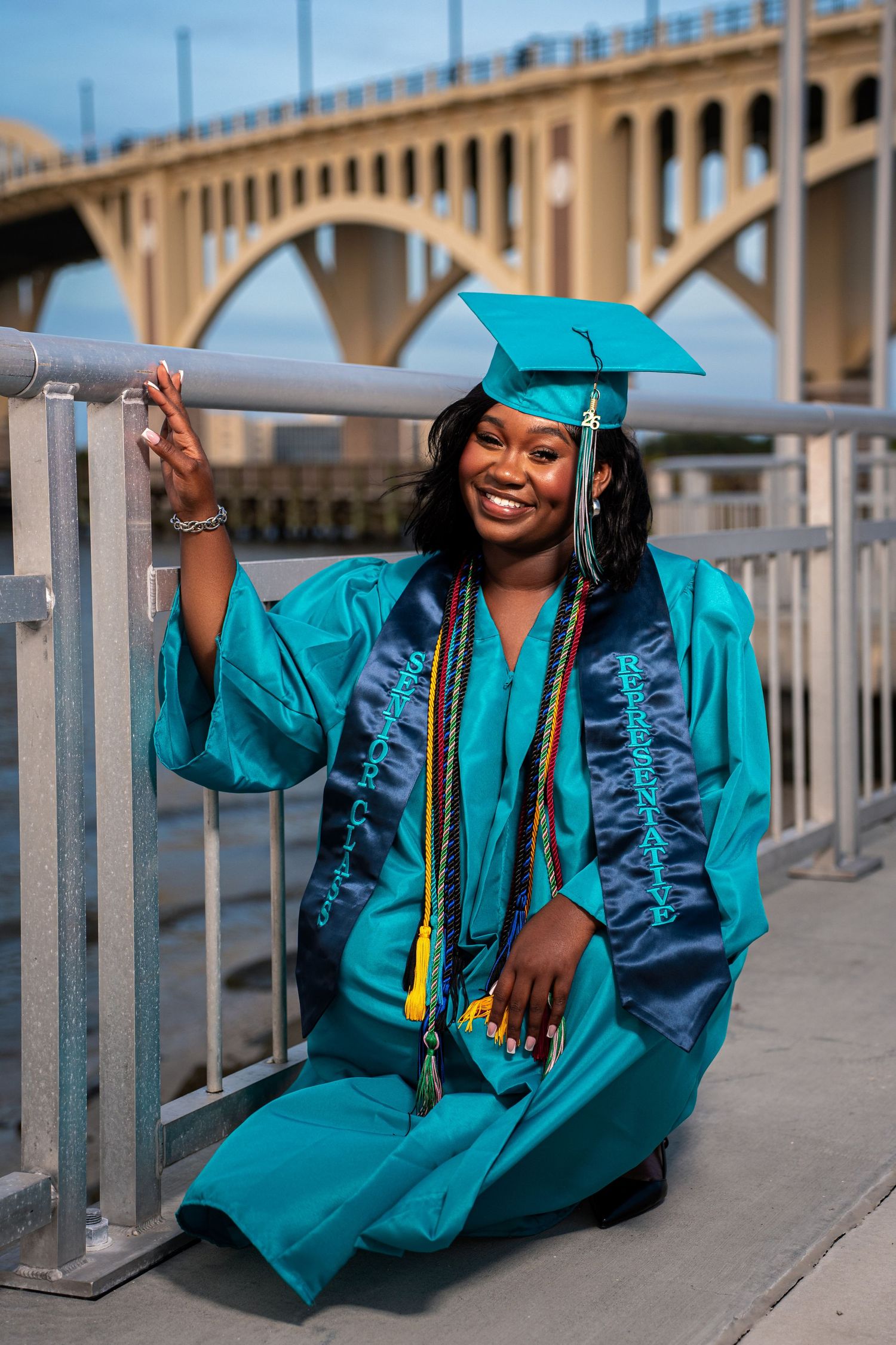 Smiling Black female graduate in teal cap and gown kneeling by railing with bridge in background.