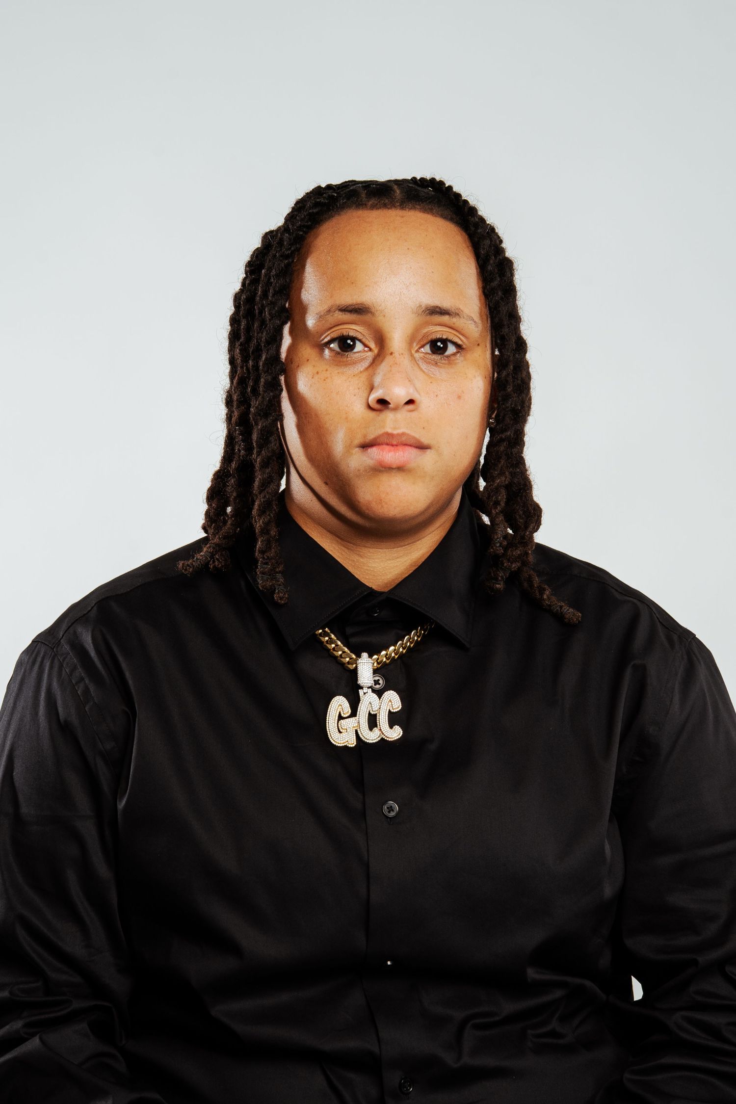 Person with dreadlocks wearing black shirt and GCC gold chain necklace against gray background.