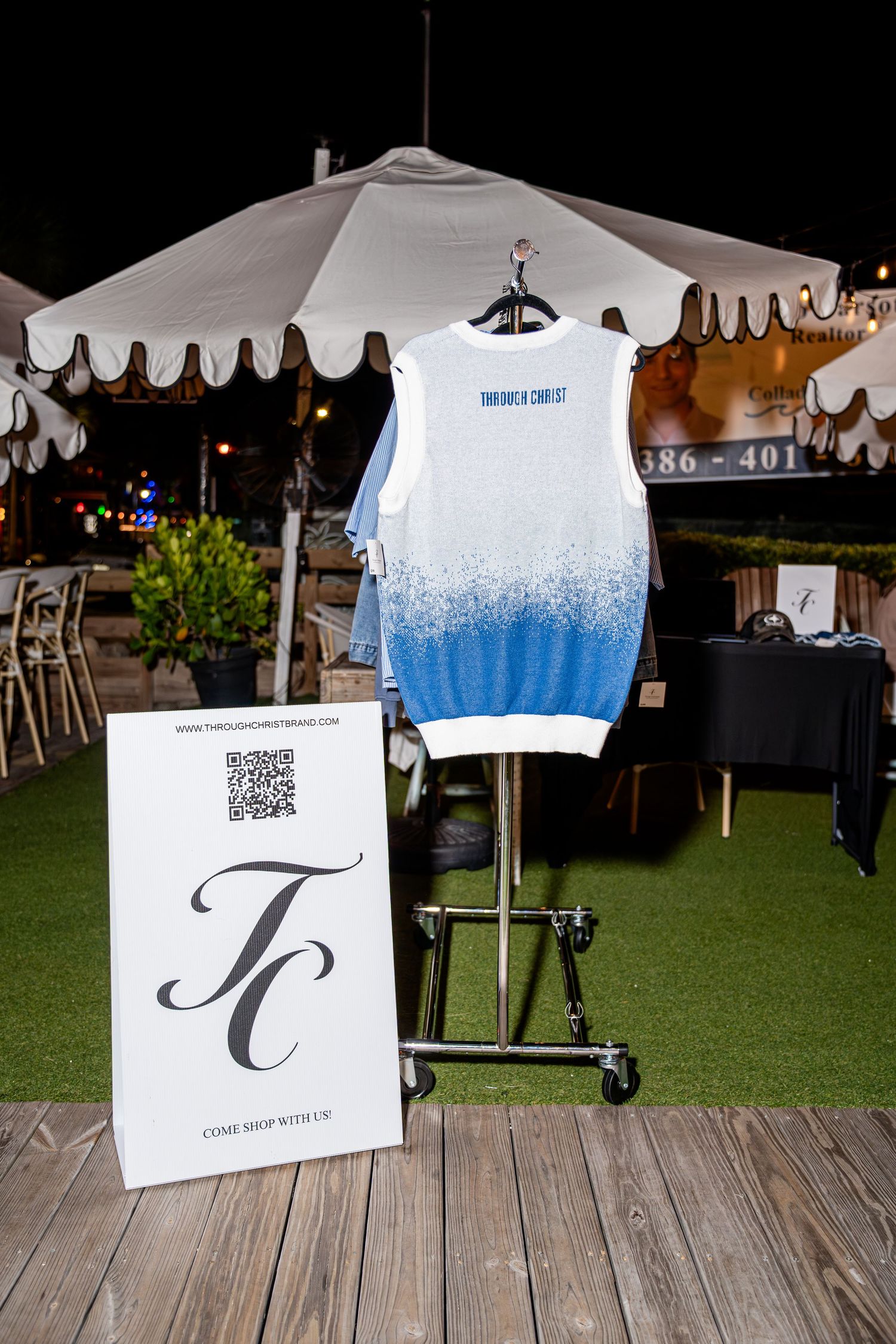 White and blue ombre sleeveless shirt displayed on a rack beside a TC brand sign at an outdoor market.
