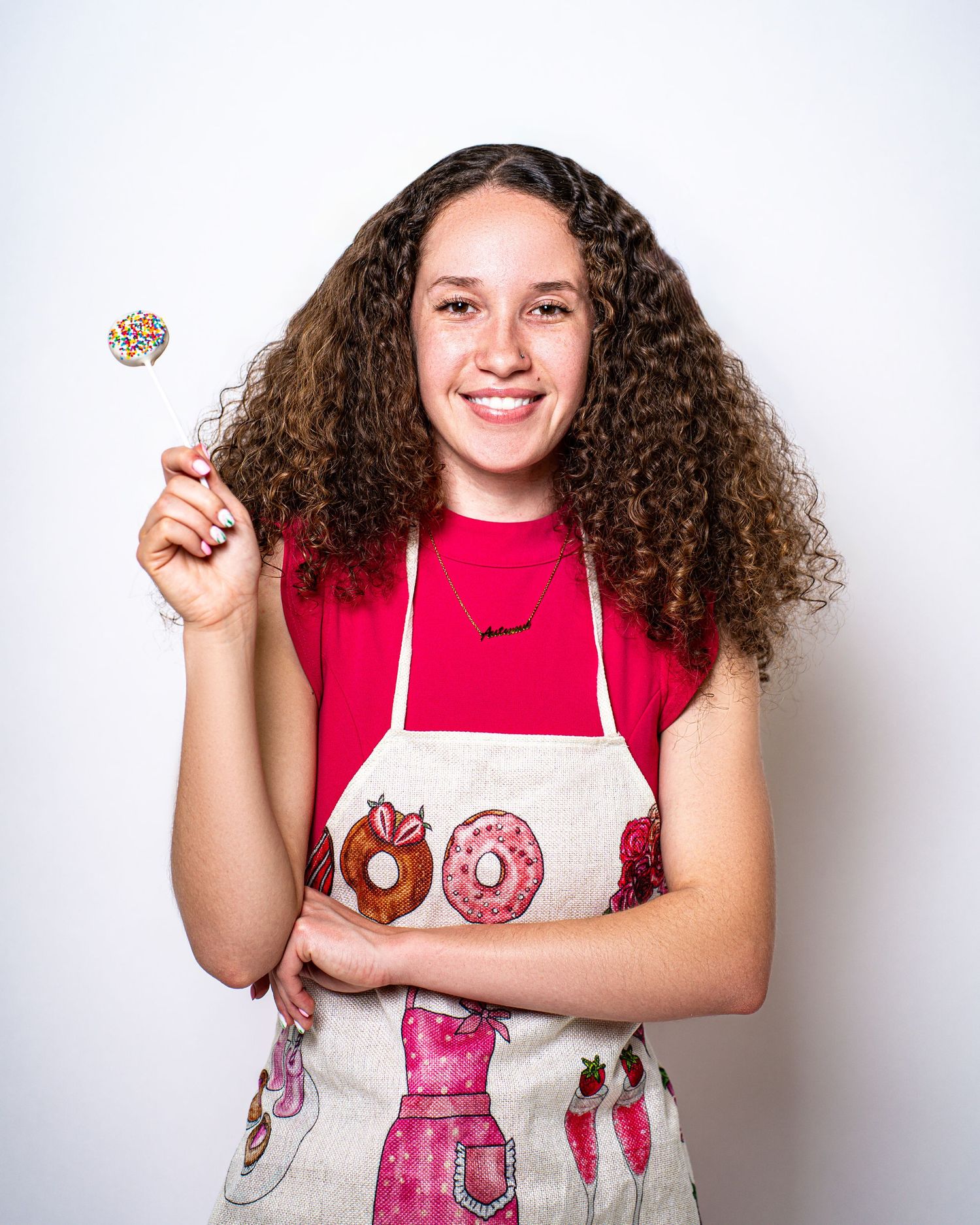 Smiling young woman with curly hair wearing a baking apron holds a colorful sprinkle cake pop.
