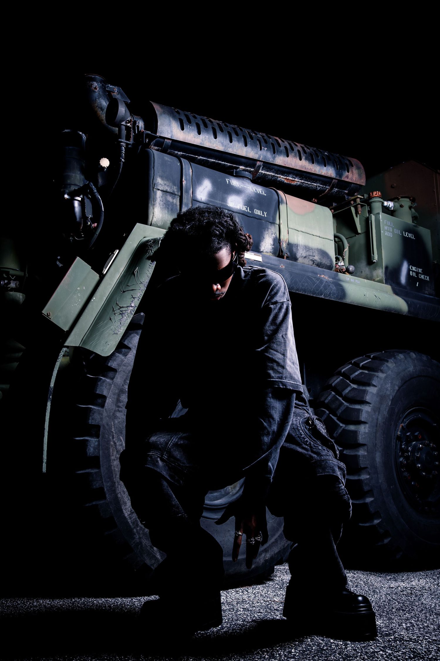 Man in dark clothing leaning against large military vehicle at night on wet pavement.