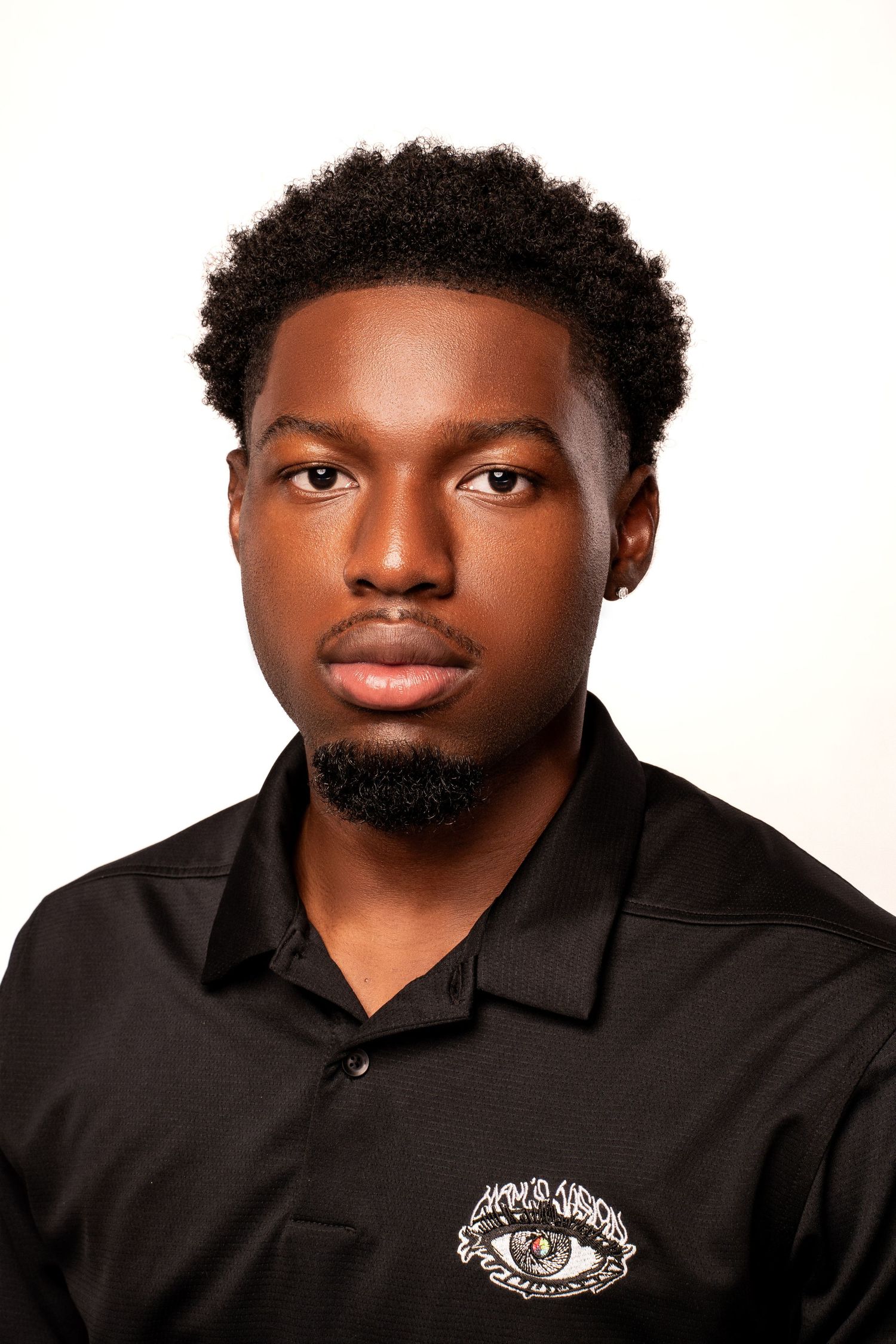 Young Black man with natural afro hair and goatee in a black branded polo shirt on white background.