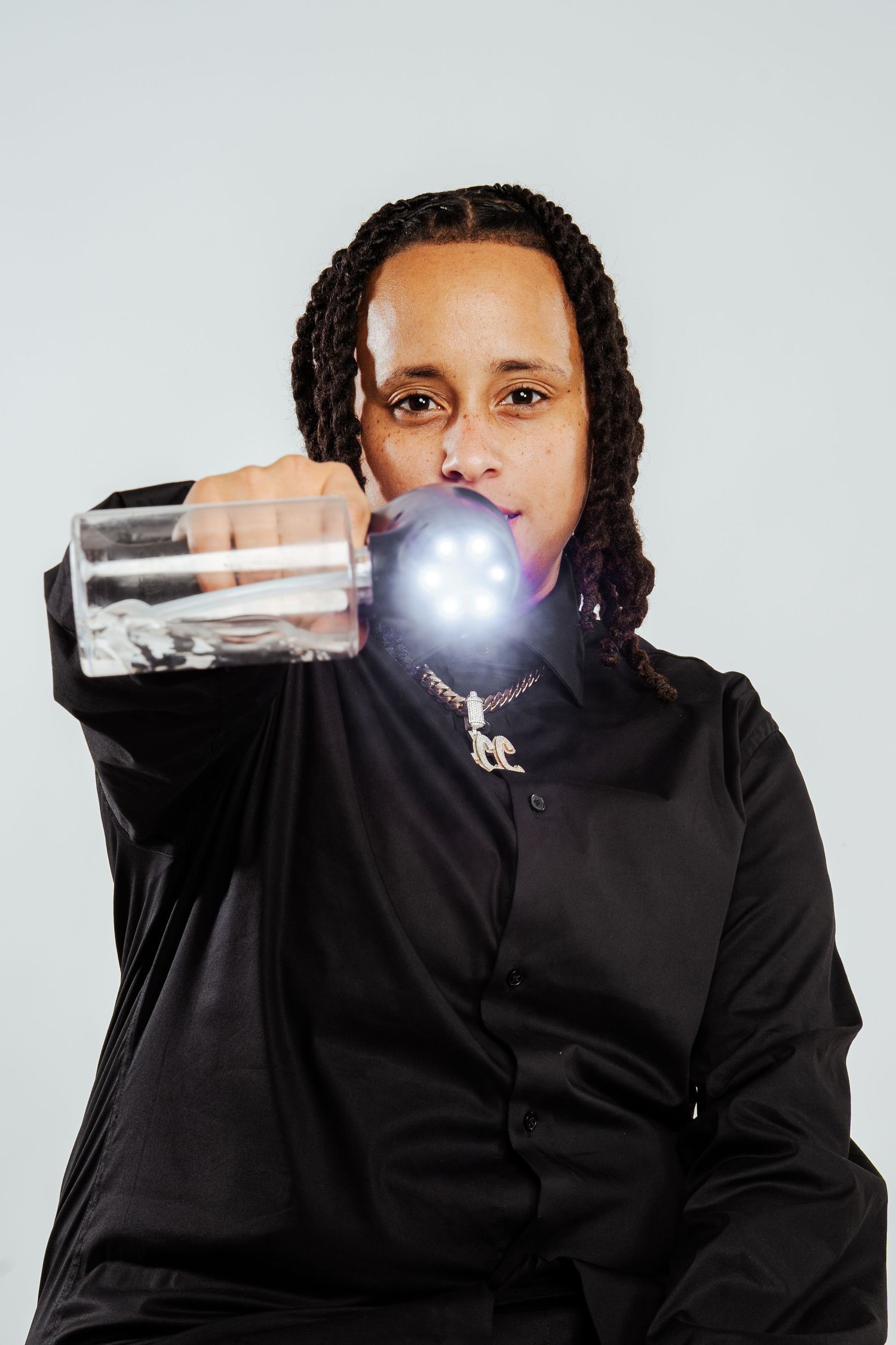 Person in black shirt holding illuminated glass bottle toward camera with chain necklace on light background.