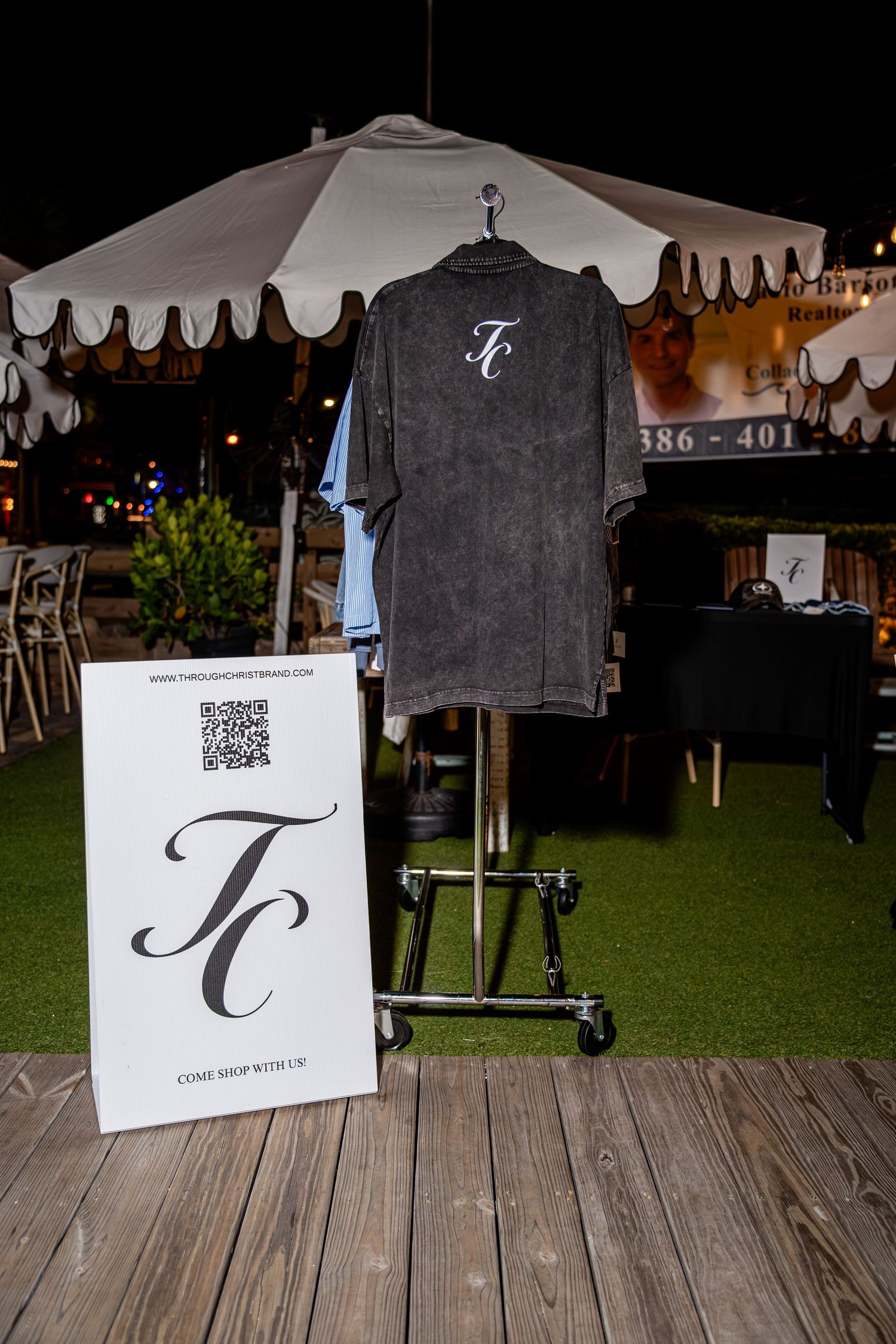 Dark gray TC monogram button-up shirt displayed on rack with TC brand QR code sign at night.