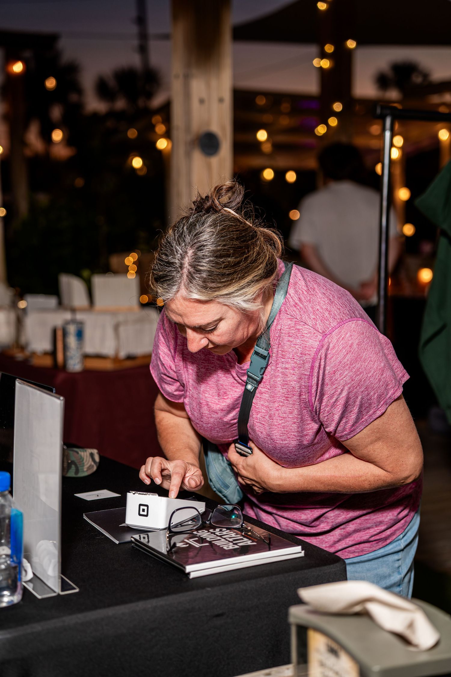Woman browsing merchandise at an outdoor vendor table with string lights in background.