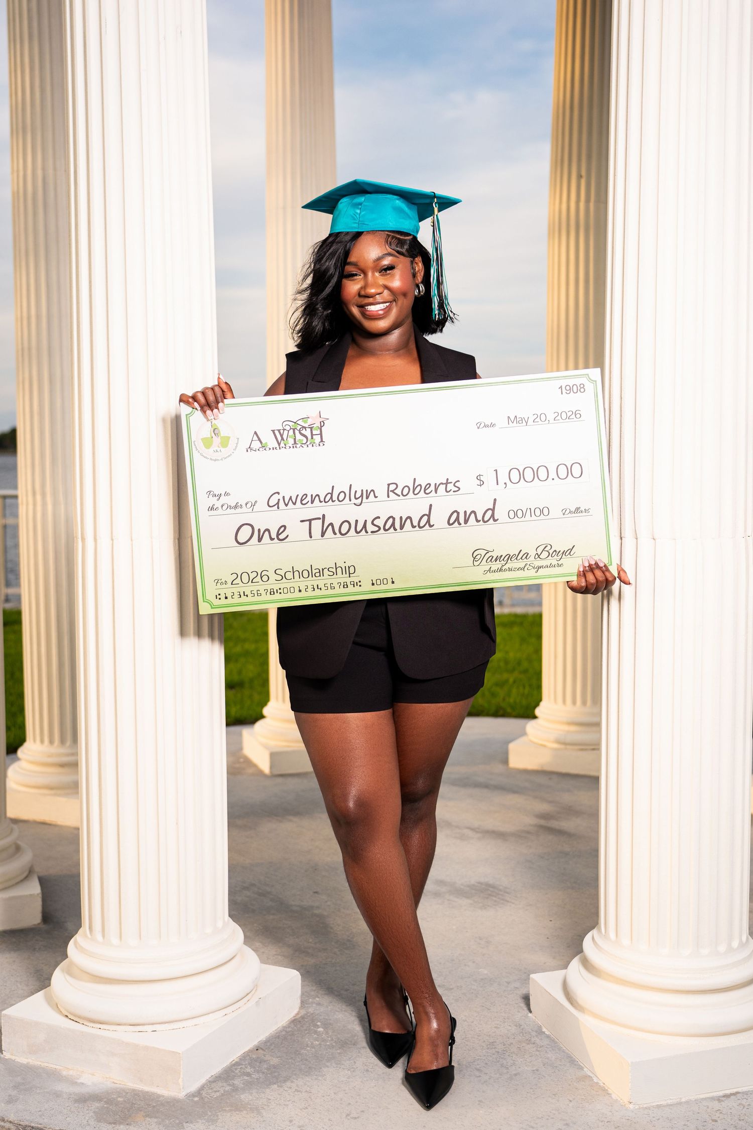Gwendolyn Roberts smiling while holding $1,000 scholarship check in teal grad cap between white columns.