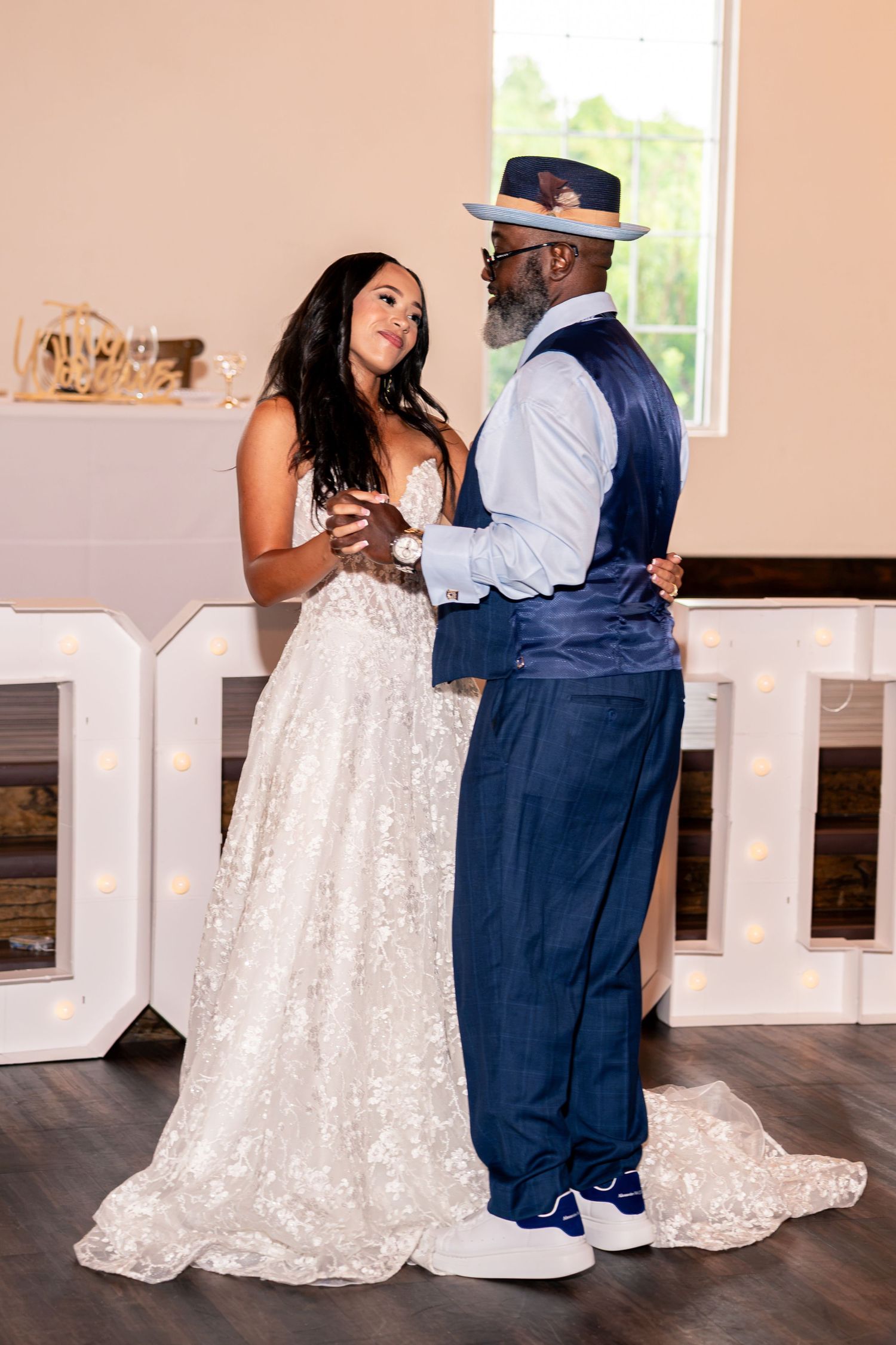 Bride in lace gown shares a tender dance with older man in navy vest and hat during wedding reception.