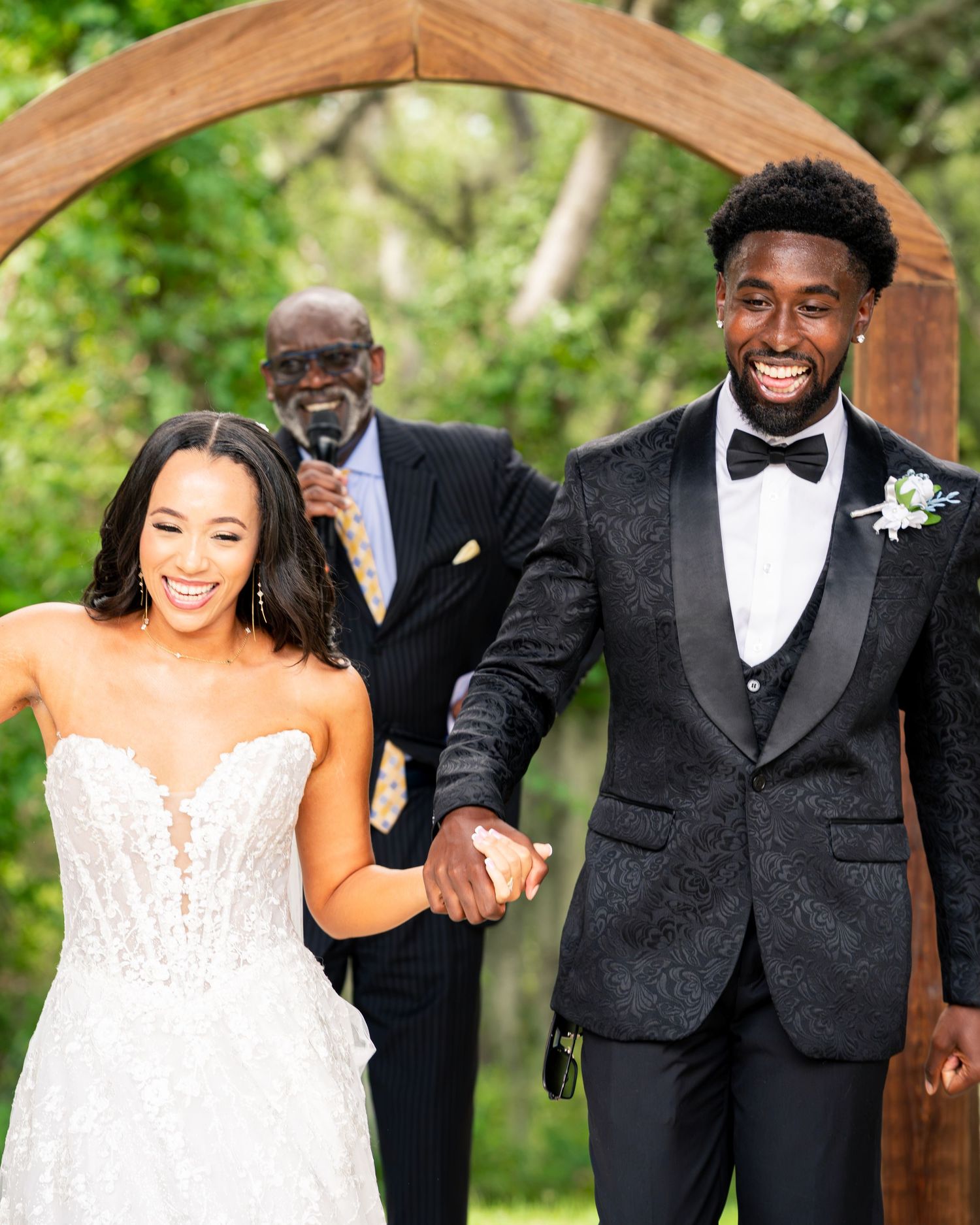 Joyful newlywed couple holding hands walking down the aisle under a wooden arch at an outdoor wedding ceremony.