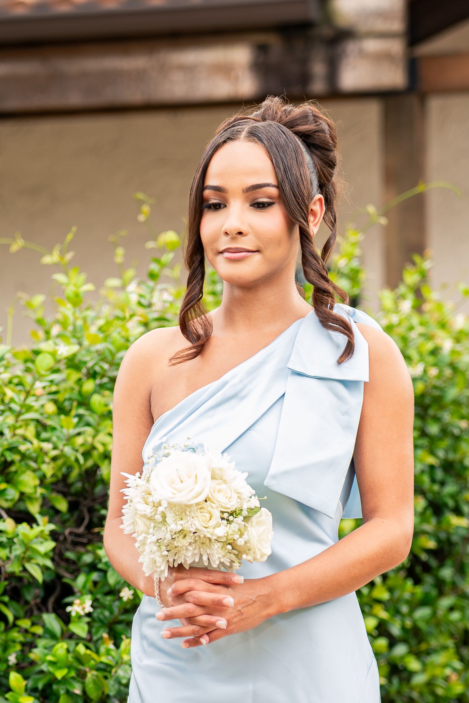 Bridesmaid in light blue one-shoulder gown holds white rose bouquet outdoors near green bushes.