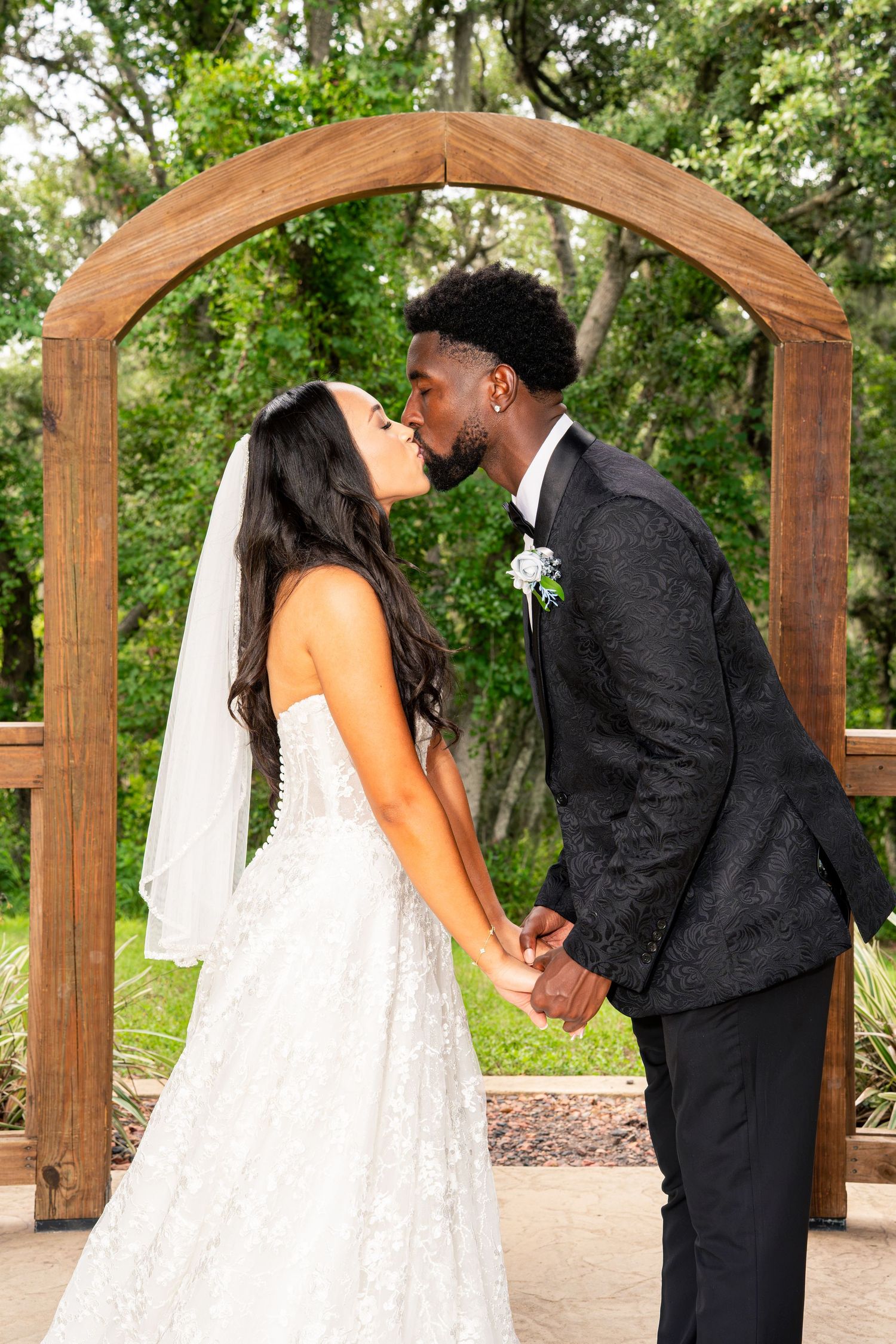 A bride and groom share a kiss under a wooden arch surrounded by lush greenery on their wedding day.