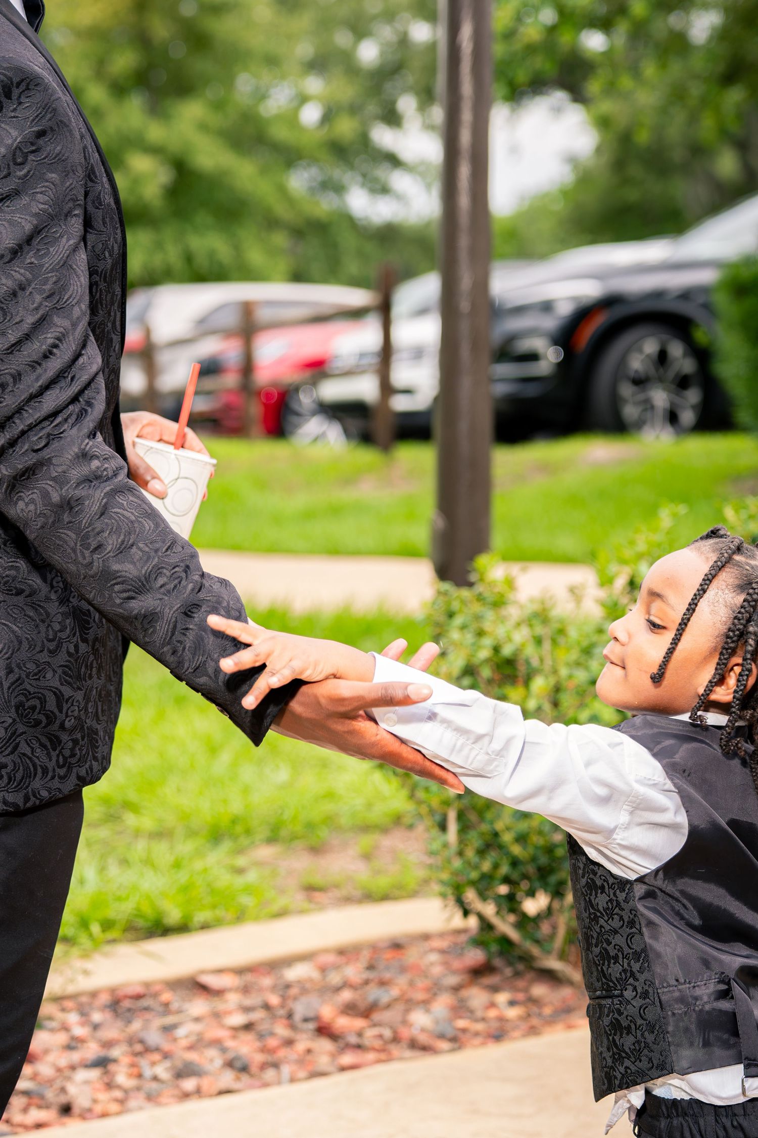 Young boy in black suit reaches up to hold hands with a wedding guest on an outdoor pathway.