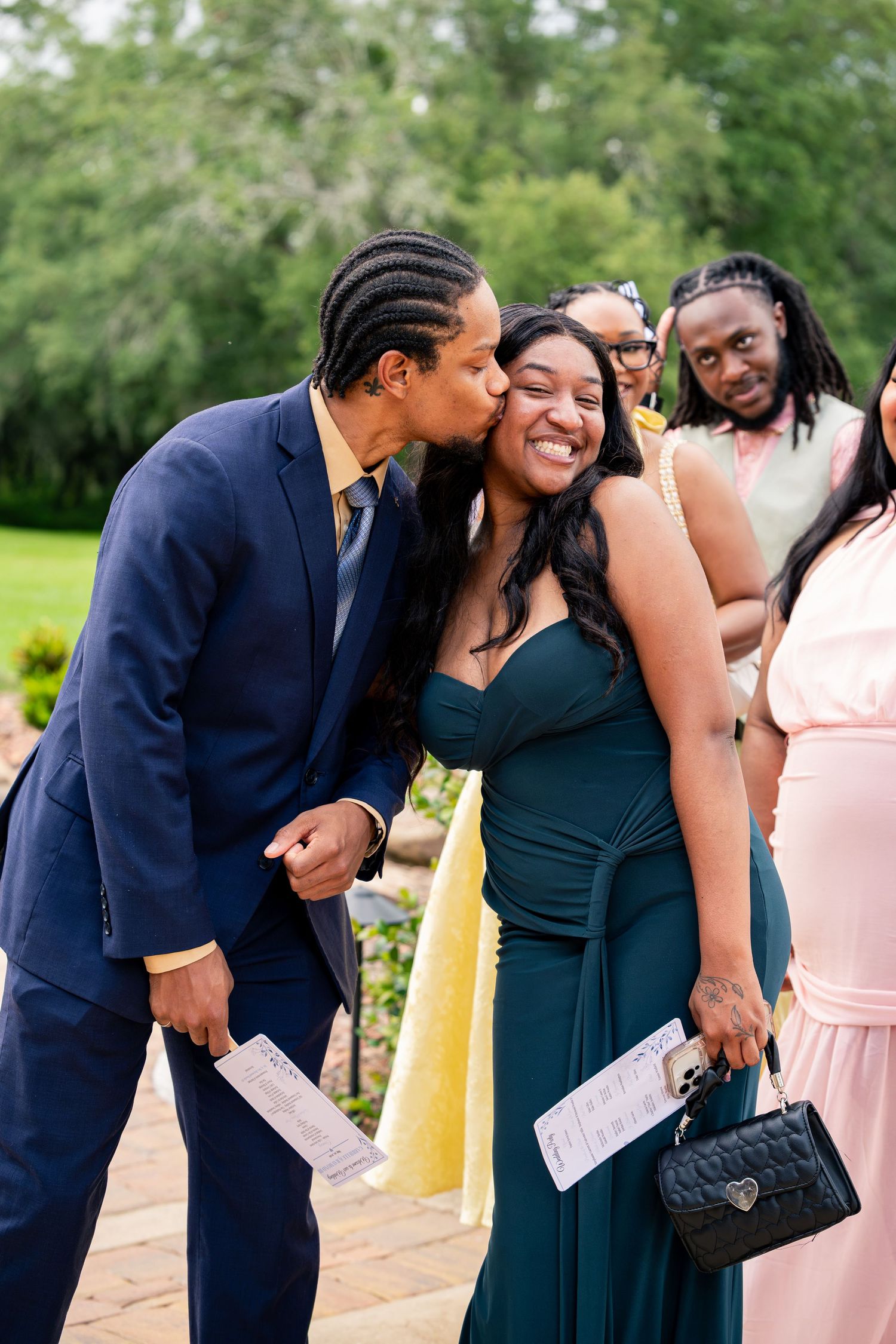 Man in navy suit kissing smiling woman in teal gown at outdoor wedding celebration.