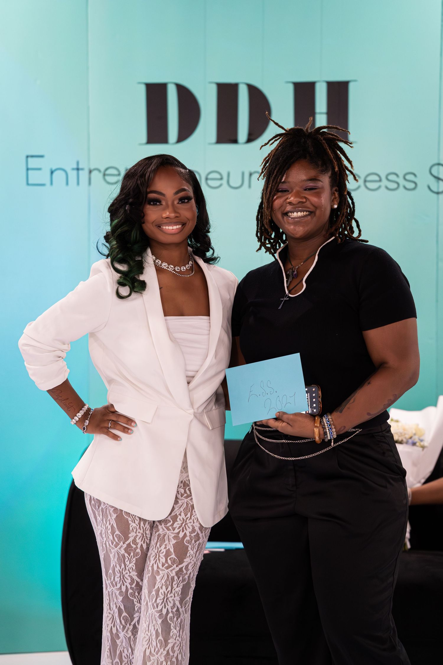 Two women smile together in front of DDH Entrepreneurial Success Summit teal backdrop.