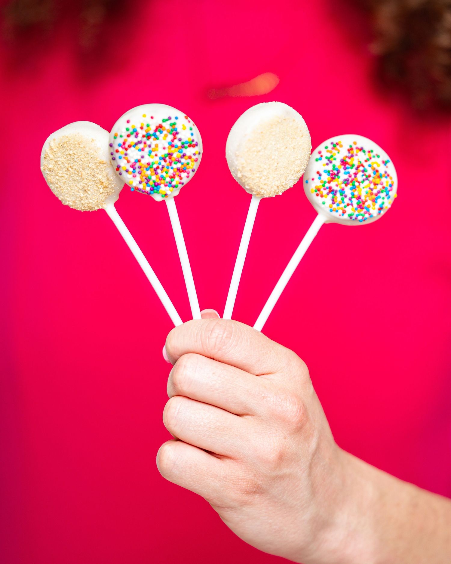 Hand holding five white chocolate cake pops with colorful sprinkles against a vibrant red background.