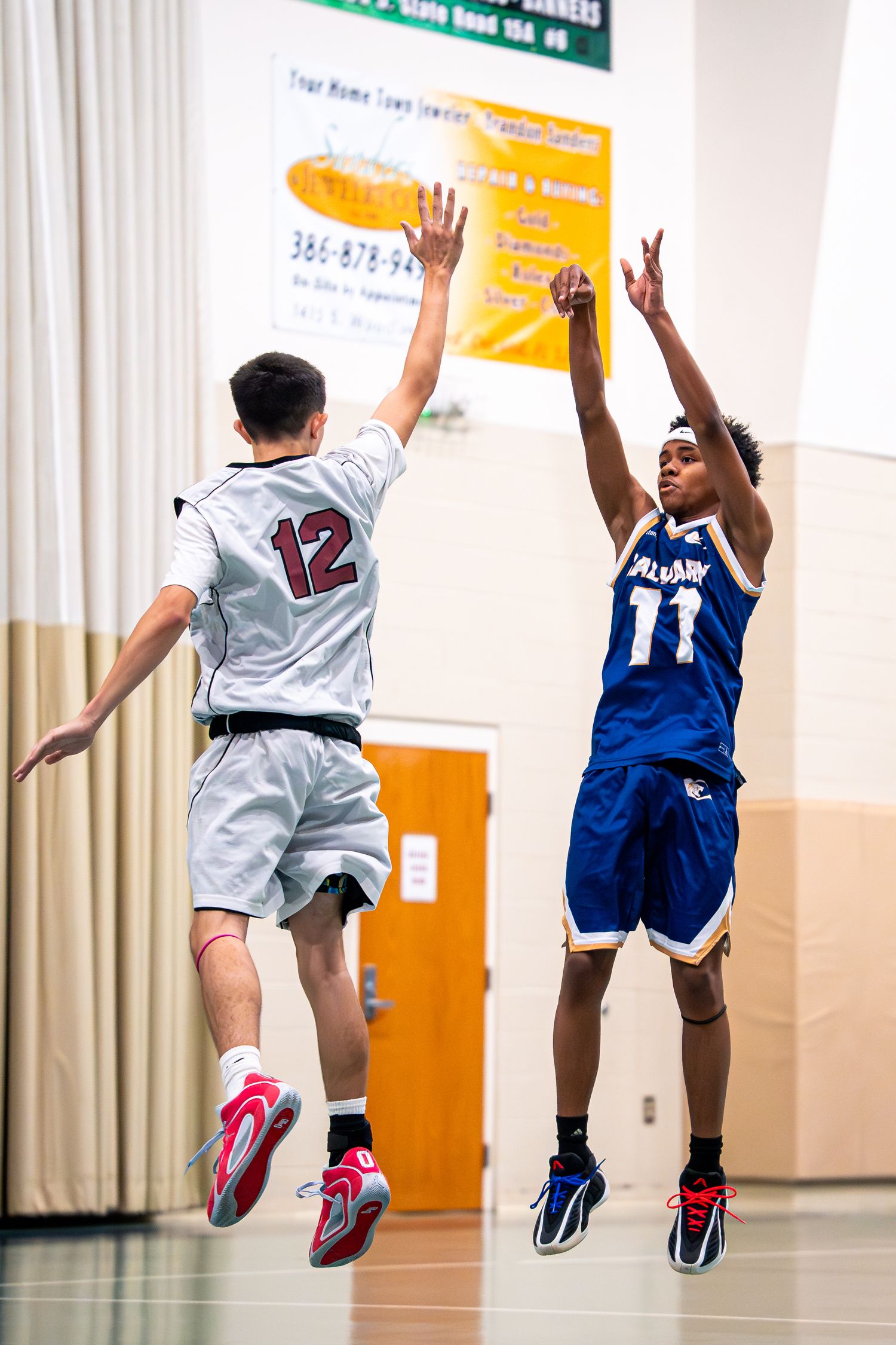 Calvary basketball player #11 shoots jump shot over defender #12 during competitive indoor game.