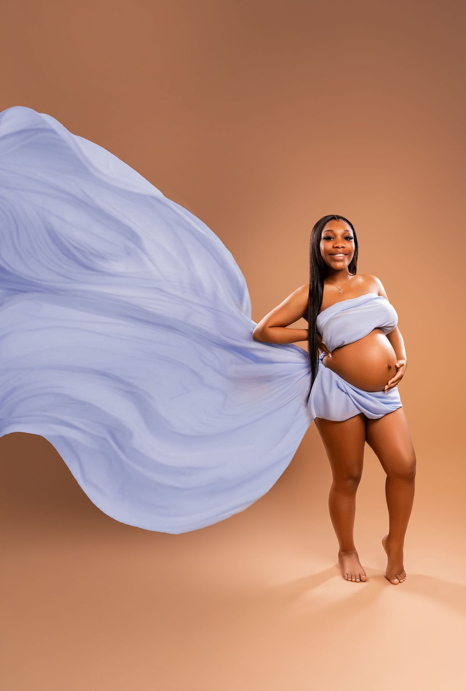 Pregnant woman in flowing blue fabric poses gracefully against warm brown background in maternity photo shoot.