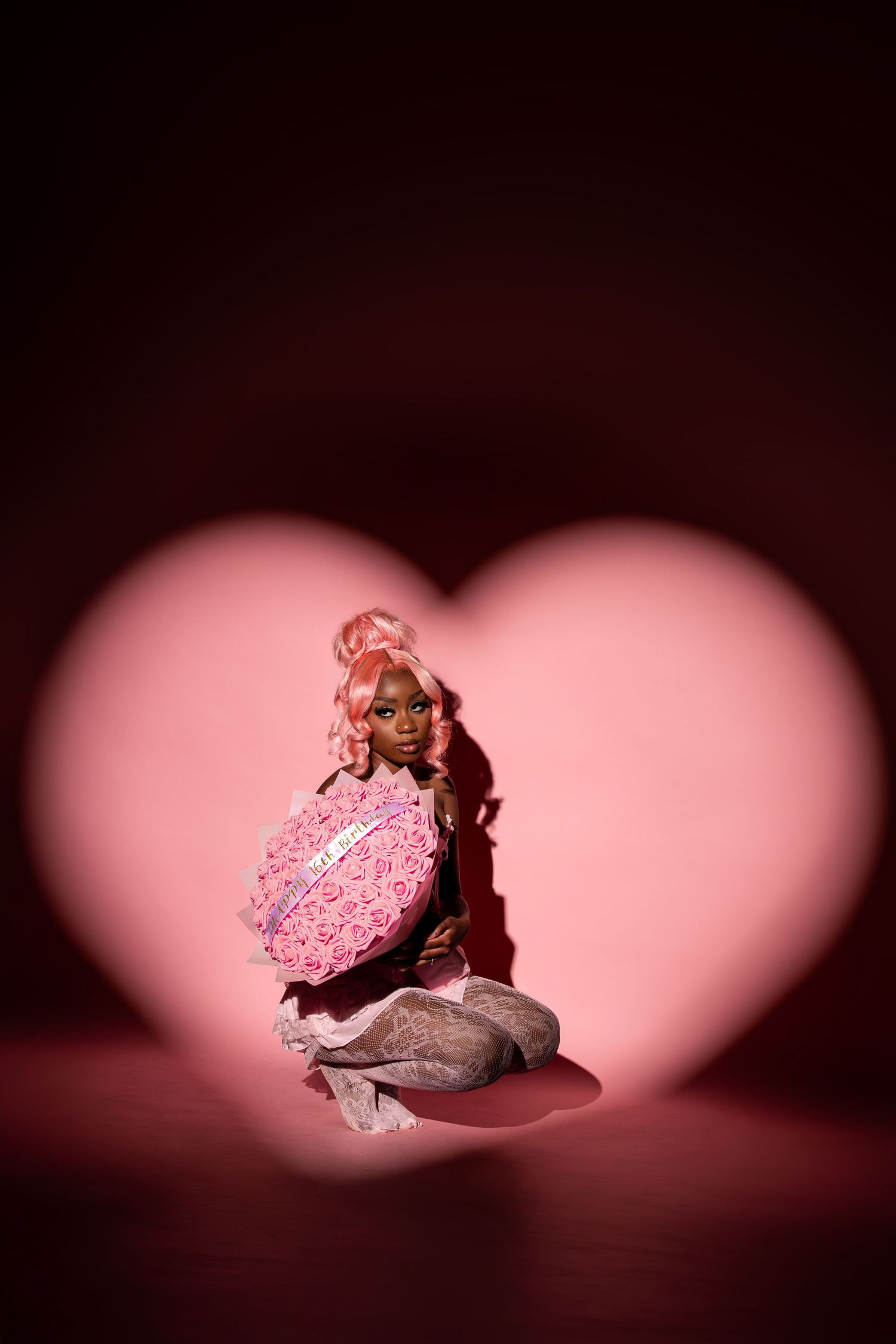 A pink-haired doll holds a heart-shaped rose box, framed by a glowing pink heart shape on a dark background.