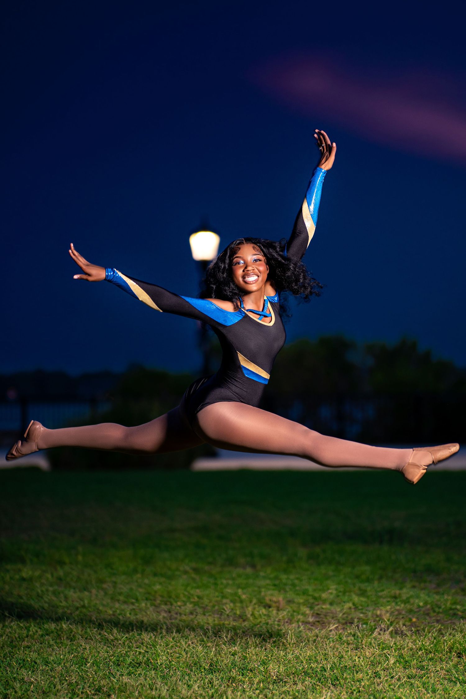 Cheerleader performing a perfect split jump in blue and black uniform on grass at night.