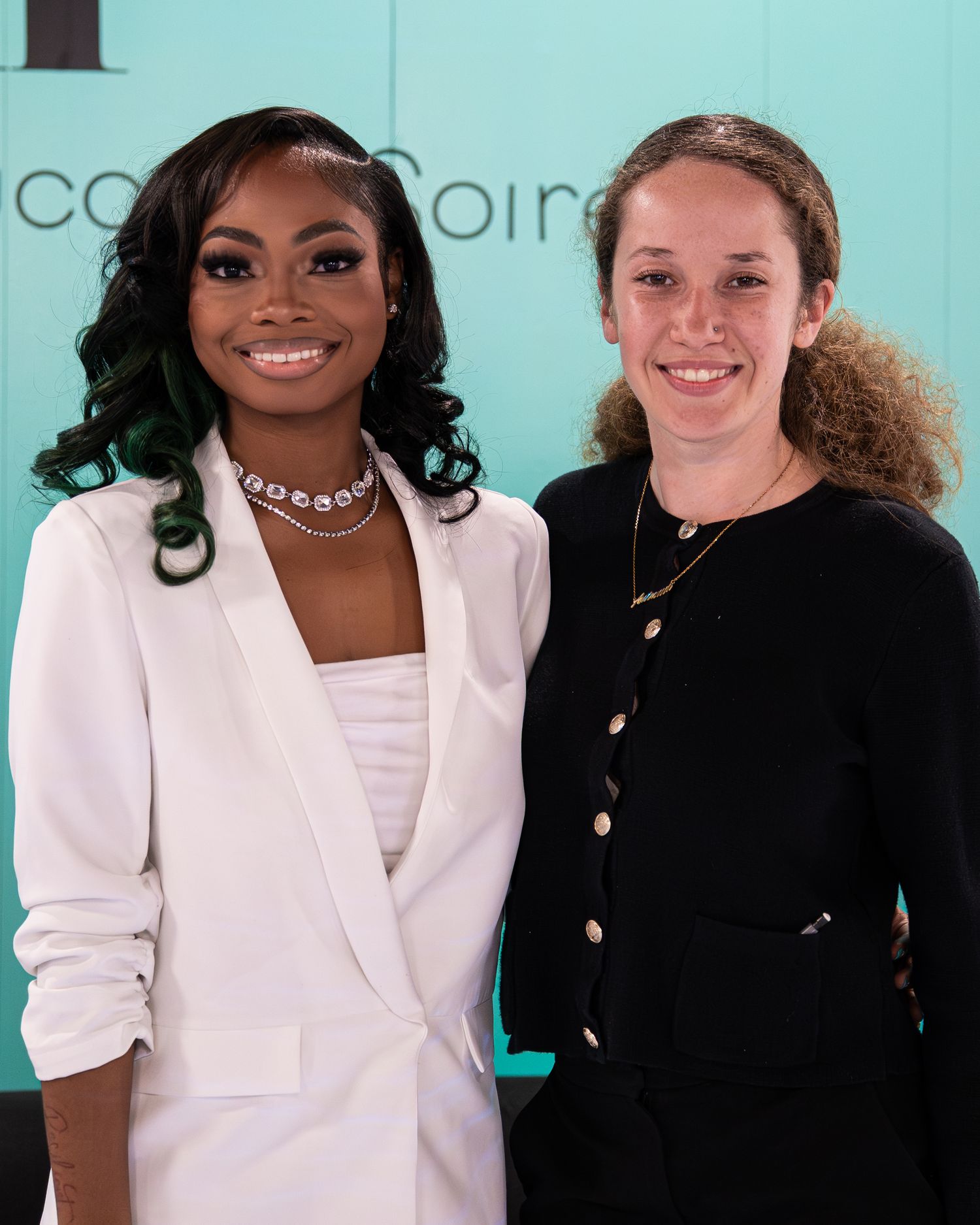 Two women smile together at DDH Entrepreneur conference, one in white blazer with green highlighted hair, other in black.