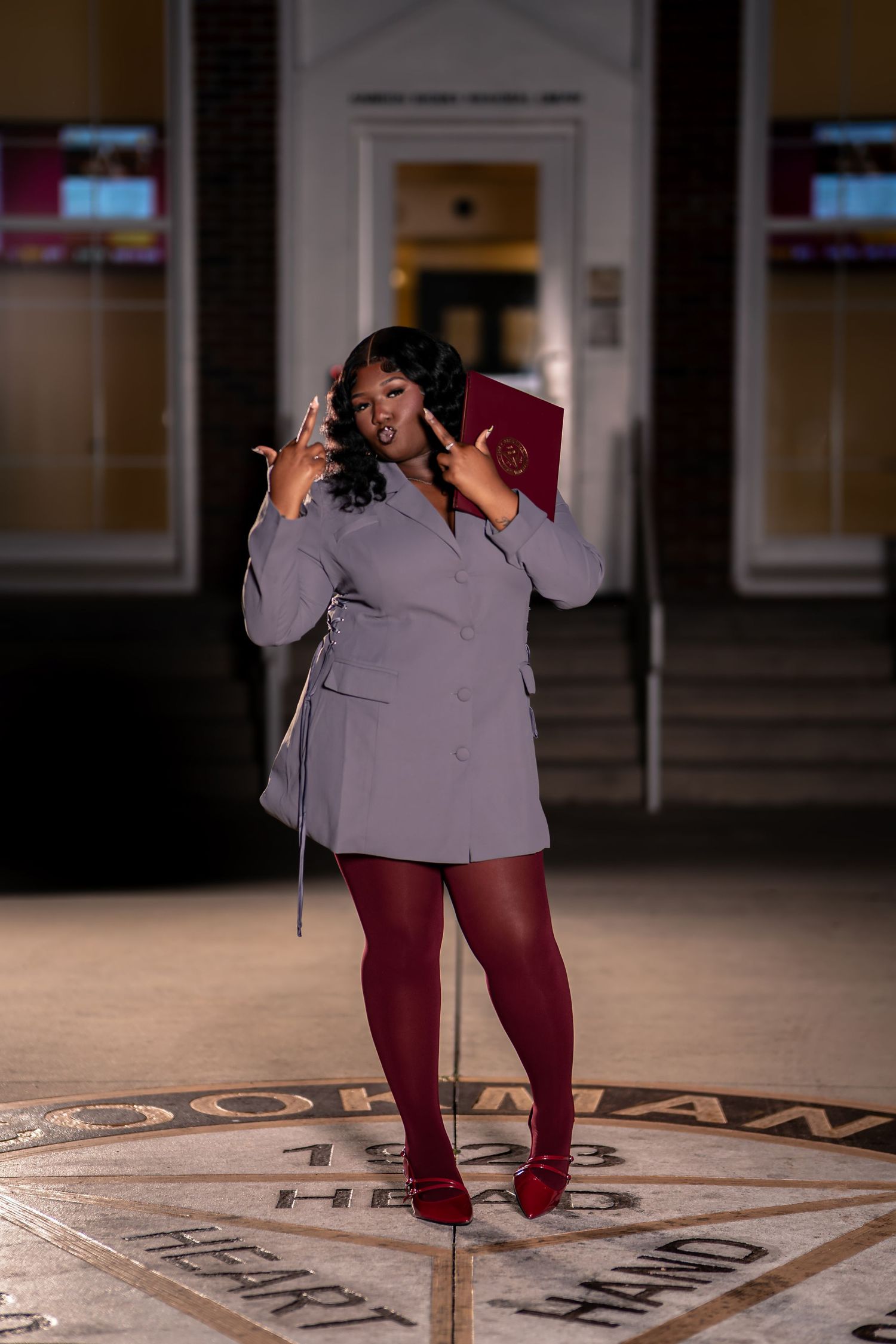 Confident graduate in gray blazer dress with maroon tights holds diploma folder on decorative floor.