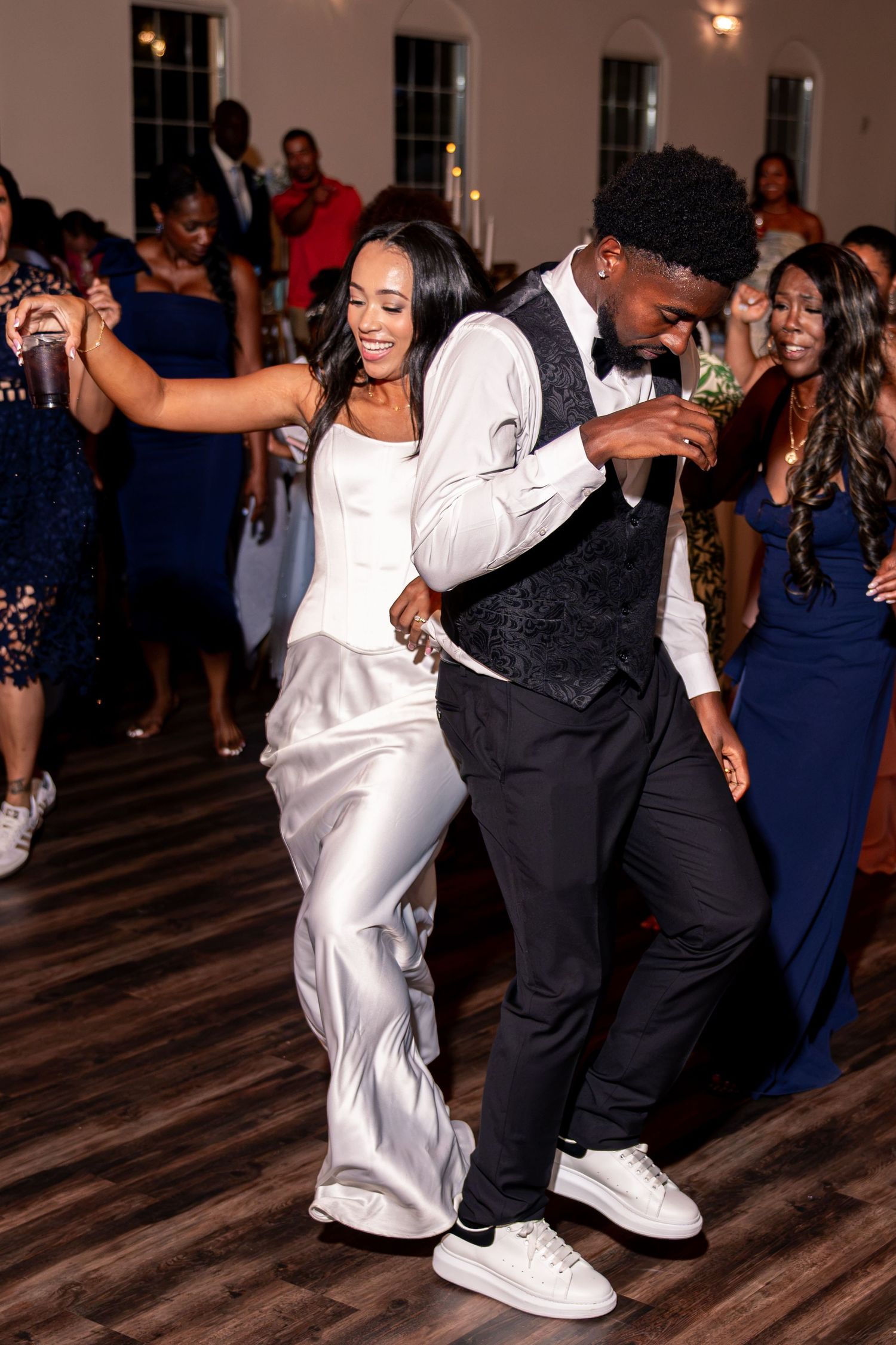 Bride in white satin gown and groom in vest dance together at lively wedding reception.