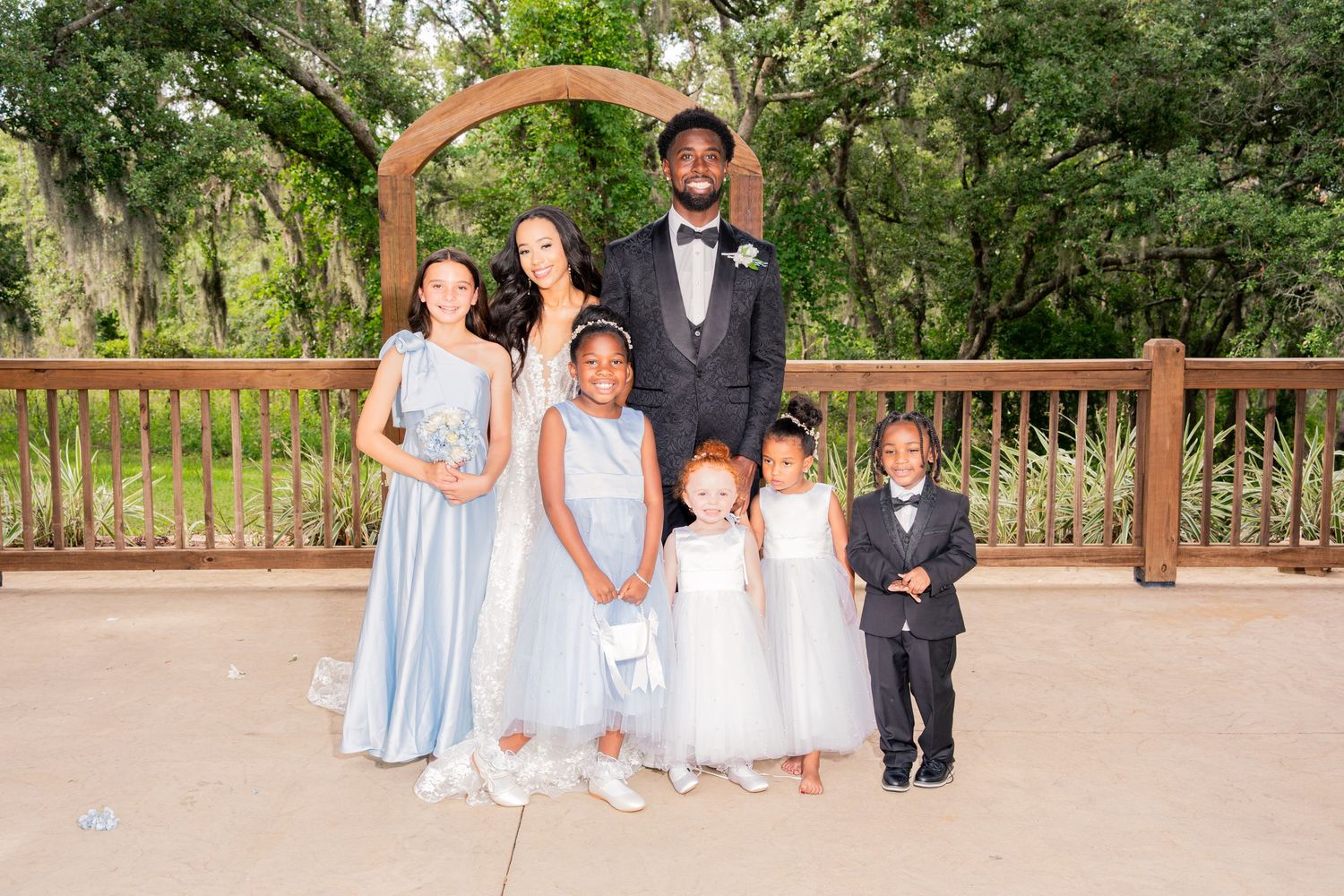 Bride and groom pose with young flower girls and ring bearer under wooden arch.